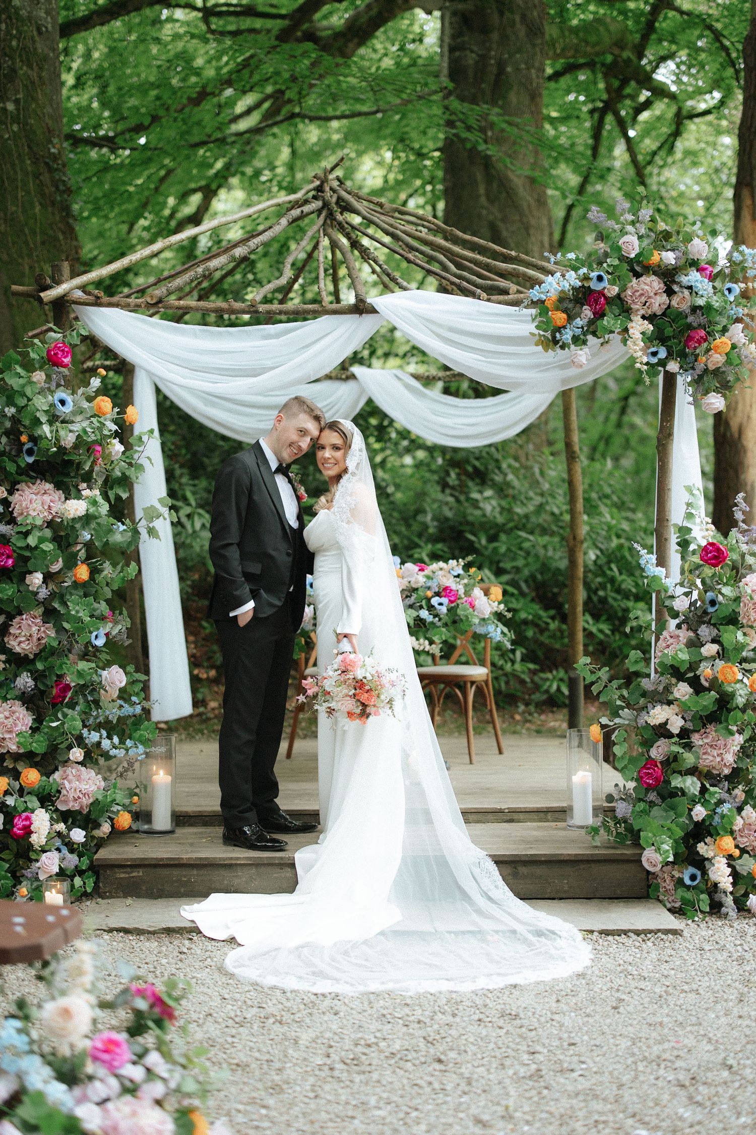 Bride and groom stand under floral arch with white draping in lush forest wedding ceremony setting.