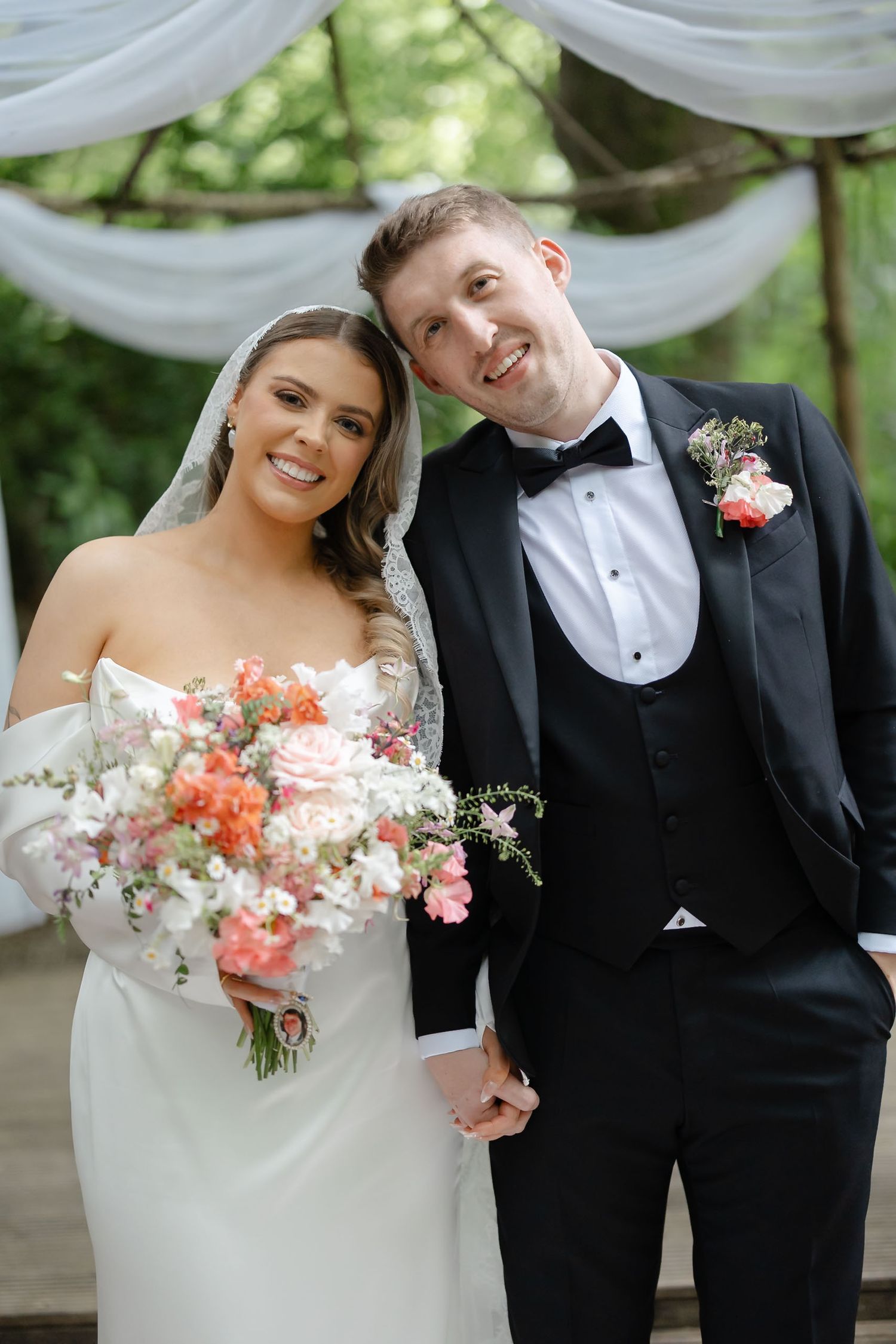 Bride and groom smile together outdoors, she holds a coral and white bouquet, he wears a black tuxedo with floral boutonniere.