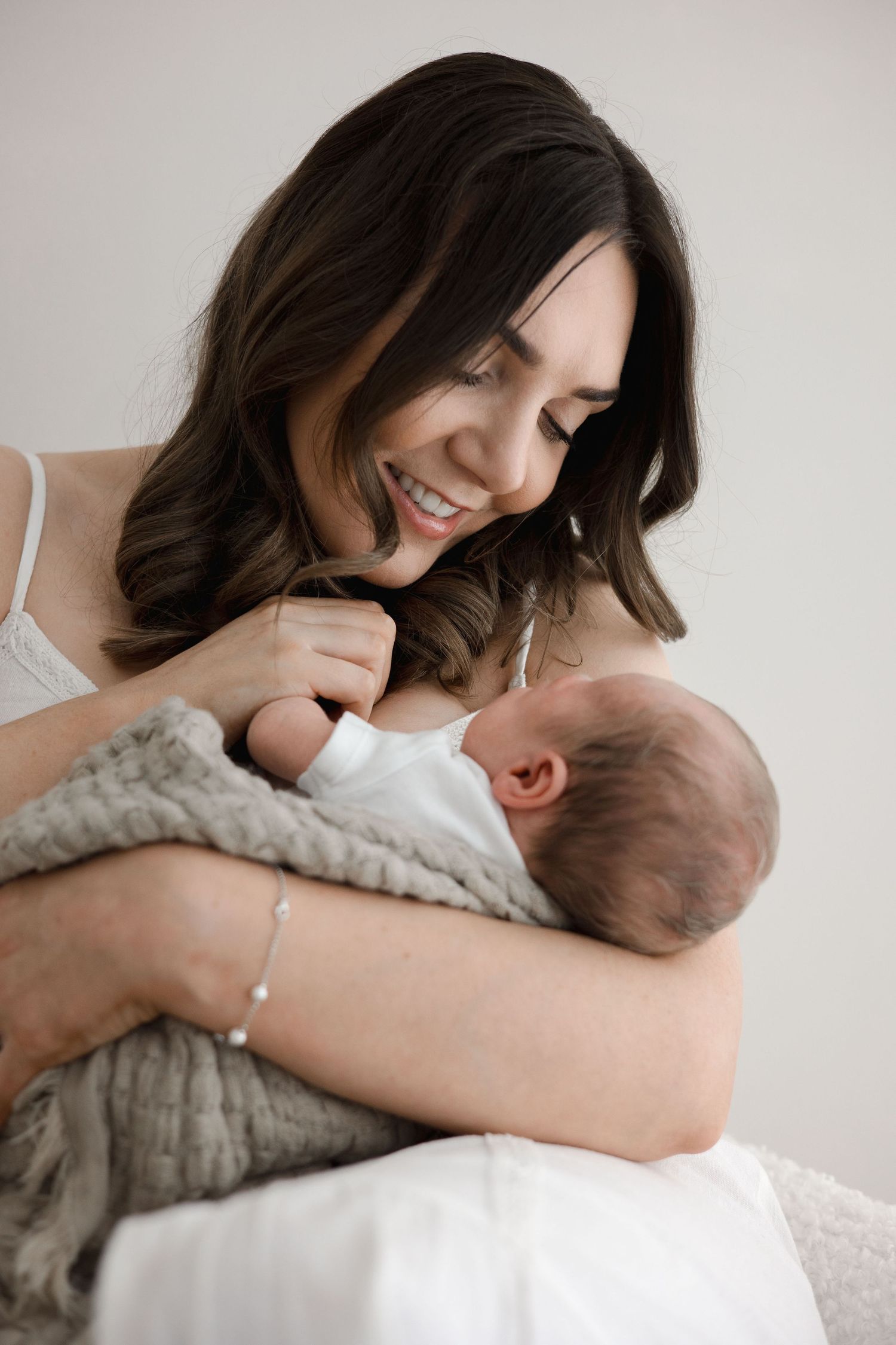 A smiling dark-haired mother holds her newborn baby wrapped in a gray blanket in a bright studio.