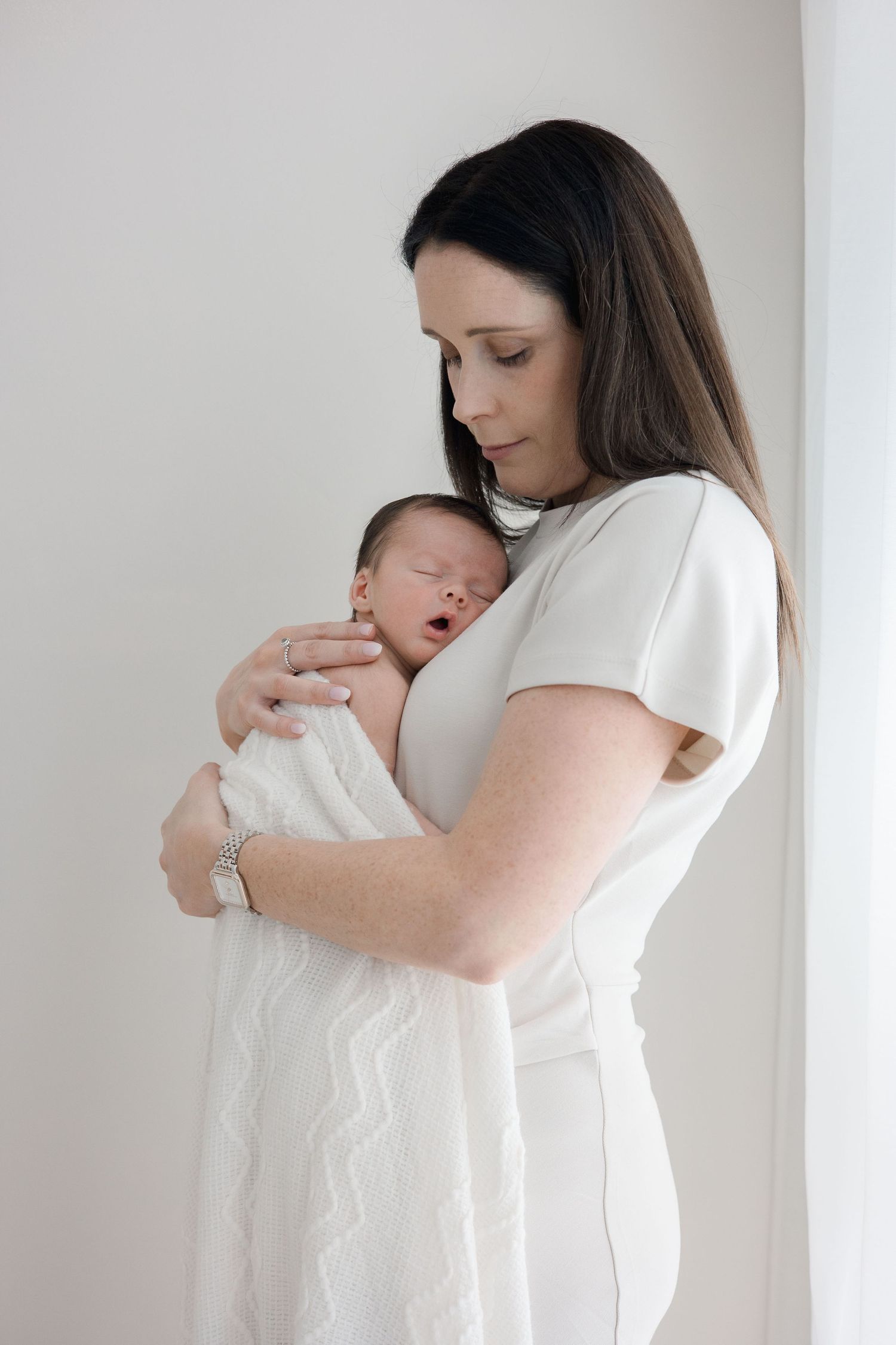 New mother in white dress gently holds sleeping swaddled newborn baby against her chest by bright window.