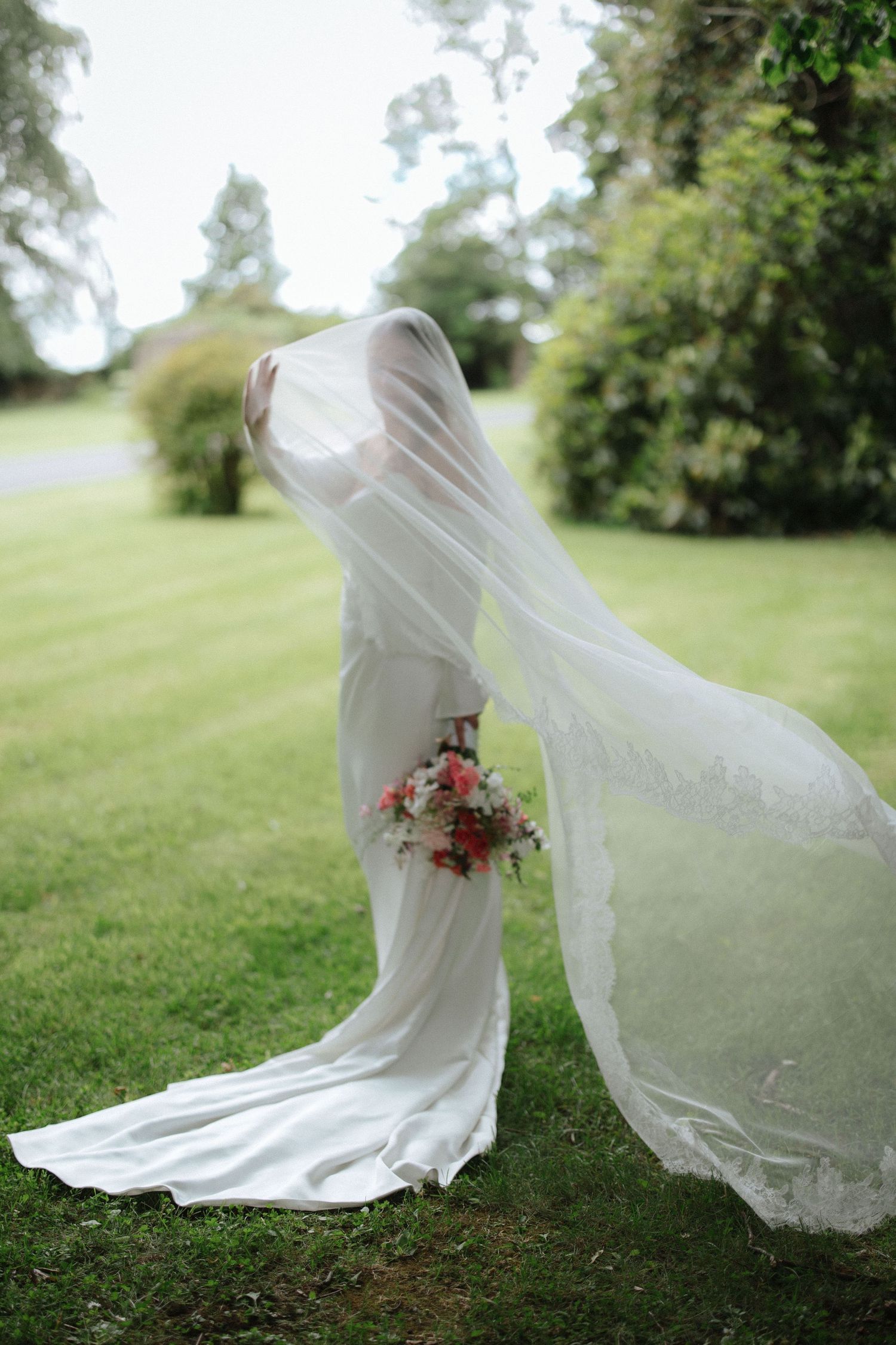 Bride in white wedding dress and flowing veil holding pink bouquet standing on green lawn with trees in background.
