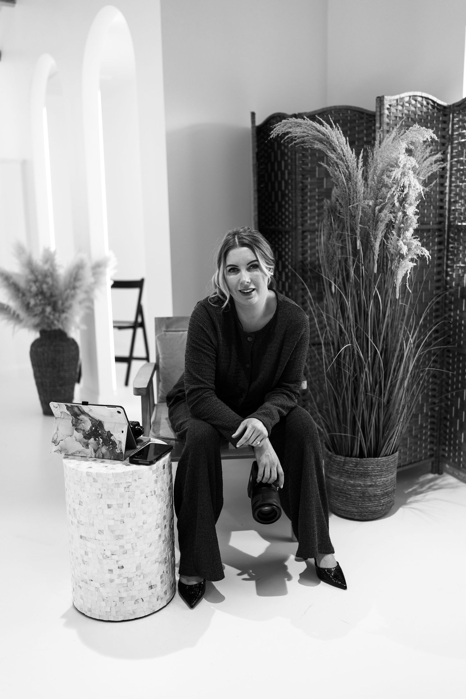 Woman in black outfit sitting with camera in stylish monochrome studio with plants and screen.