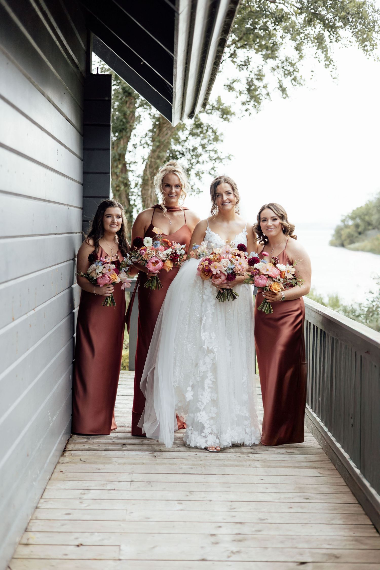 Bride in lace gown posing with three bridesmaids in rust satin dresses holding colorful bouquets.