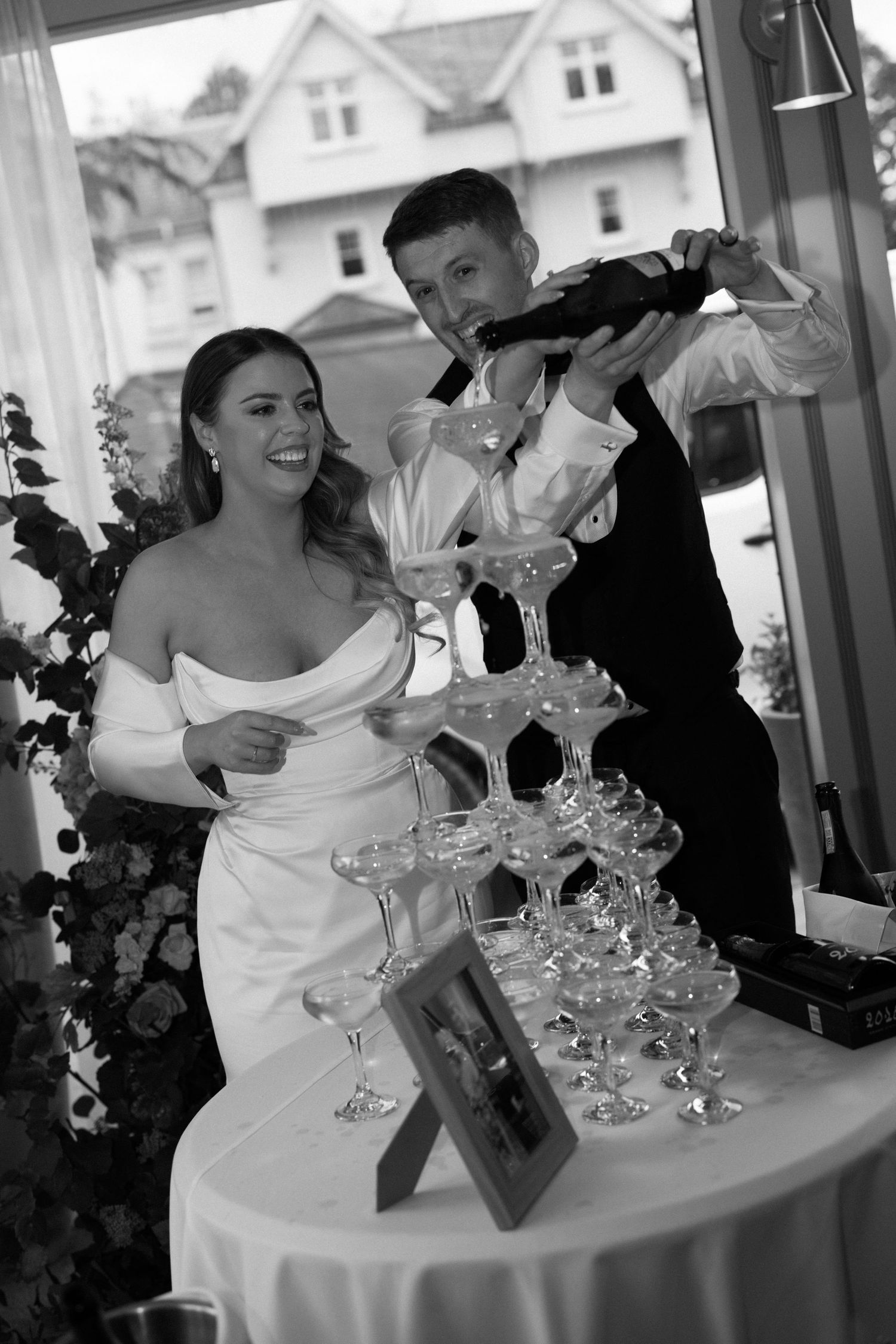 Black and white photo of bride and groom pouring champagne tower at wedding reception.