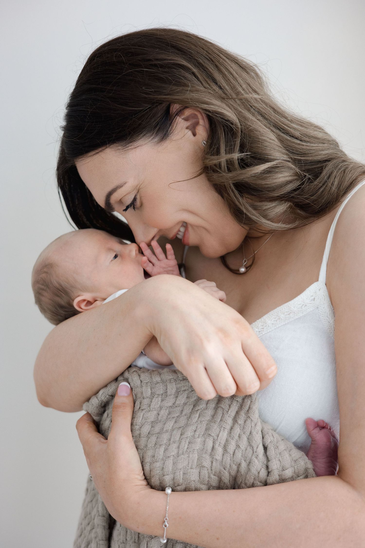 A smiling mother tenderly holds her newborn baby wrapped in a gray knit blanket, leaning in close.