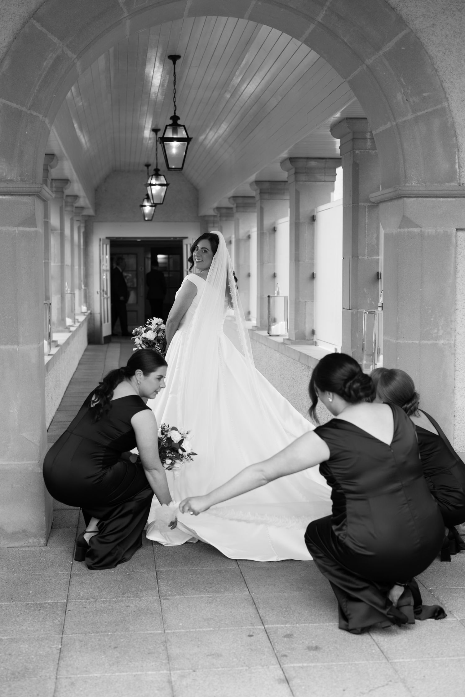 Black and white photo of a bride in a white gown assisted by two bridesmaids in a classic arched corridor.