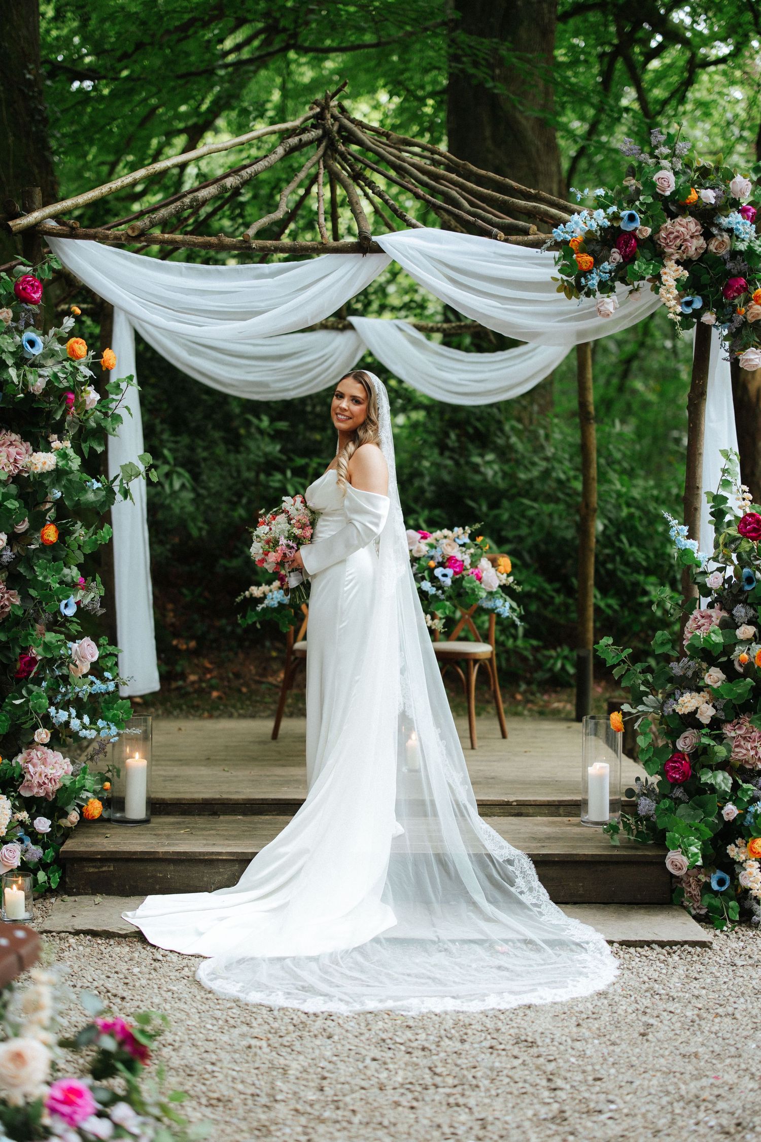Bride in white gown and veil stands under floral arch with colorful blooms and draped fabric in a lush garden setting.