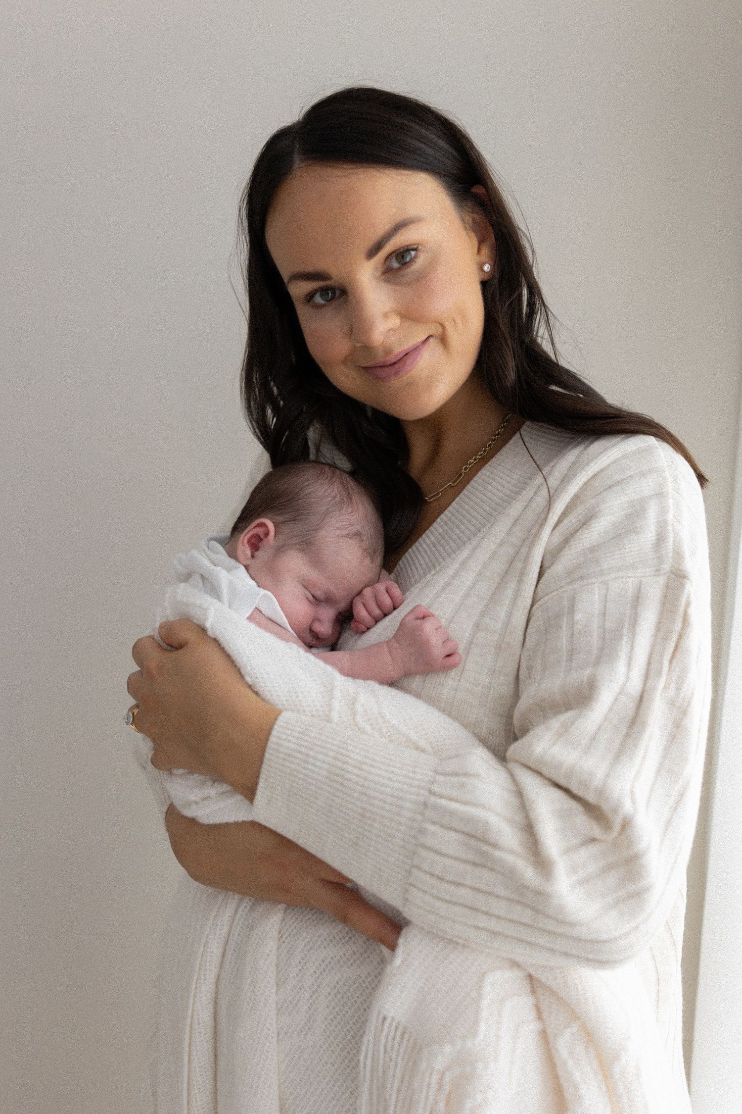 A brunette mother in a cream robe tenderly cradling her sleeping newborn baby against her chest.