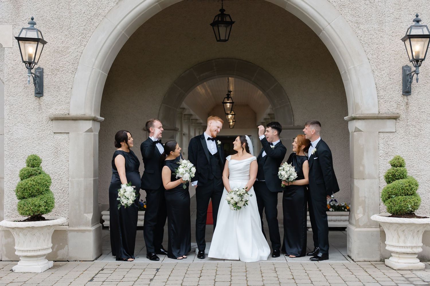 Wedding party in black attire with bride in white gown posing under stone arch at elegant venue.