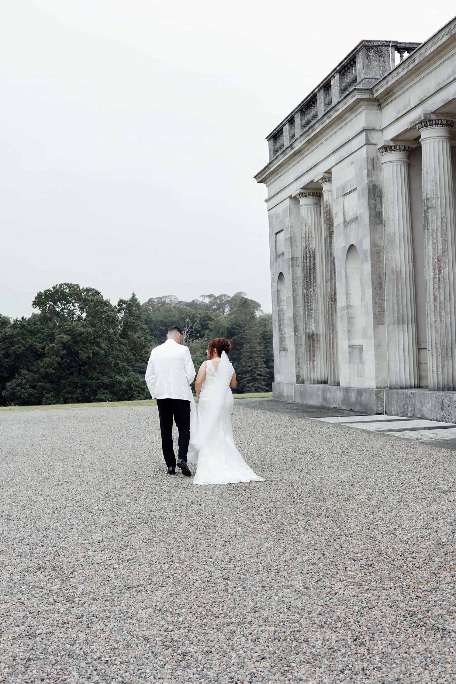 Bride and groom walking hand in hand along gravel path beside a grand neoclassical building.