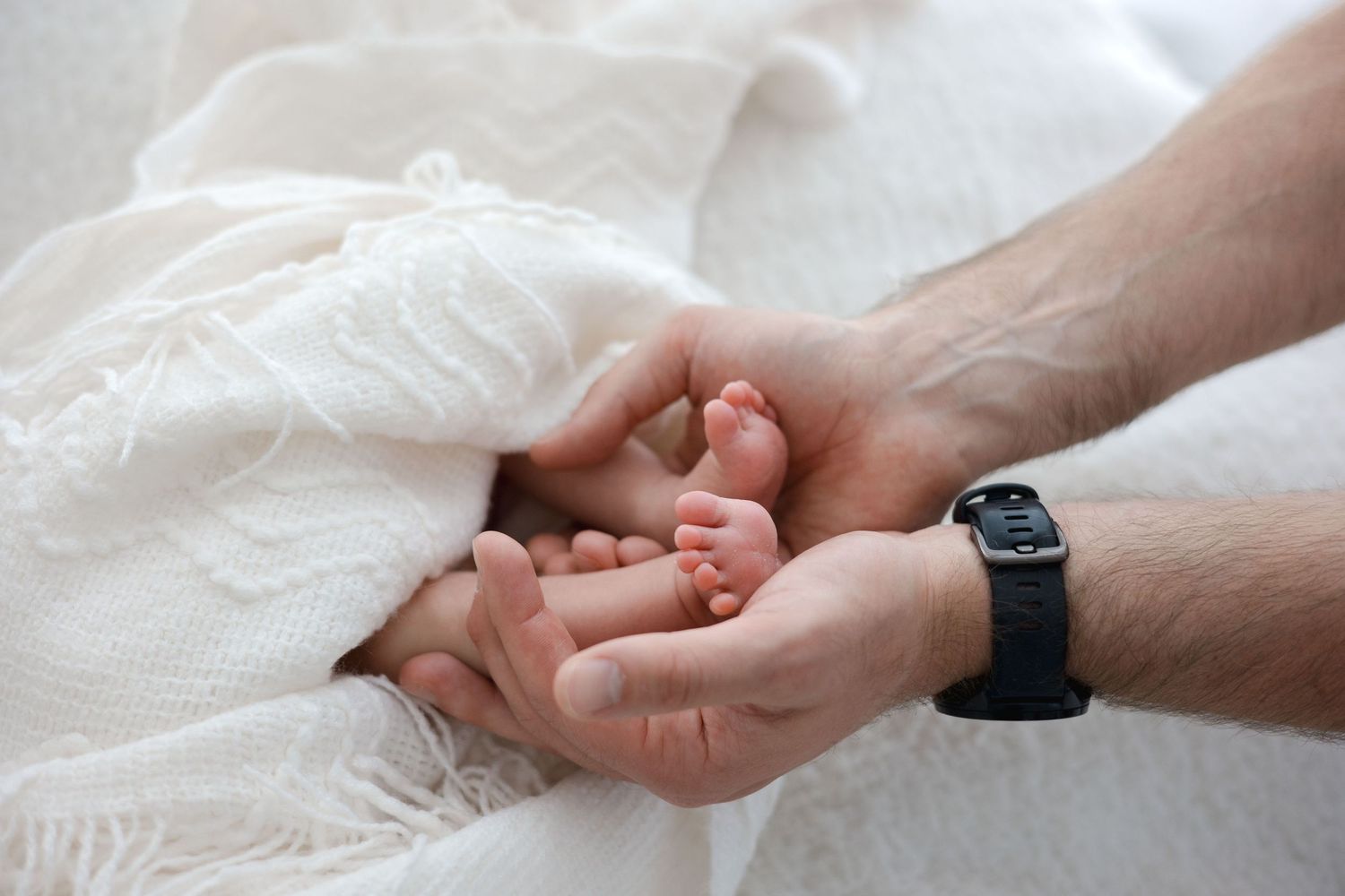 Father's hands gently cradling tiny newborn baby feet on a white blanket.