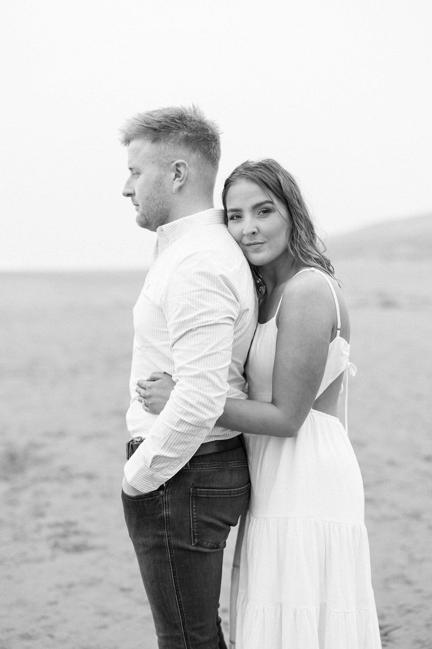 Couple embraces tenderly on misty beach in romantic black and white engagement photo, woman in white dress.