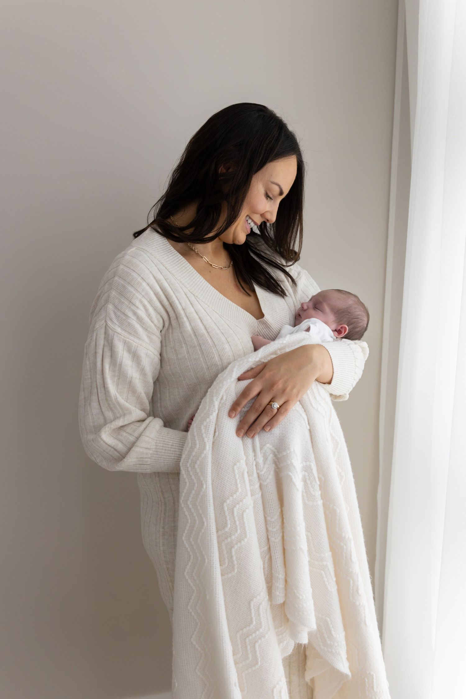 Smiling mother in cream dress holding her swaddled newborn baby by a window.