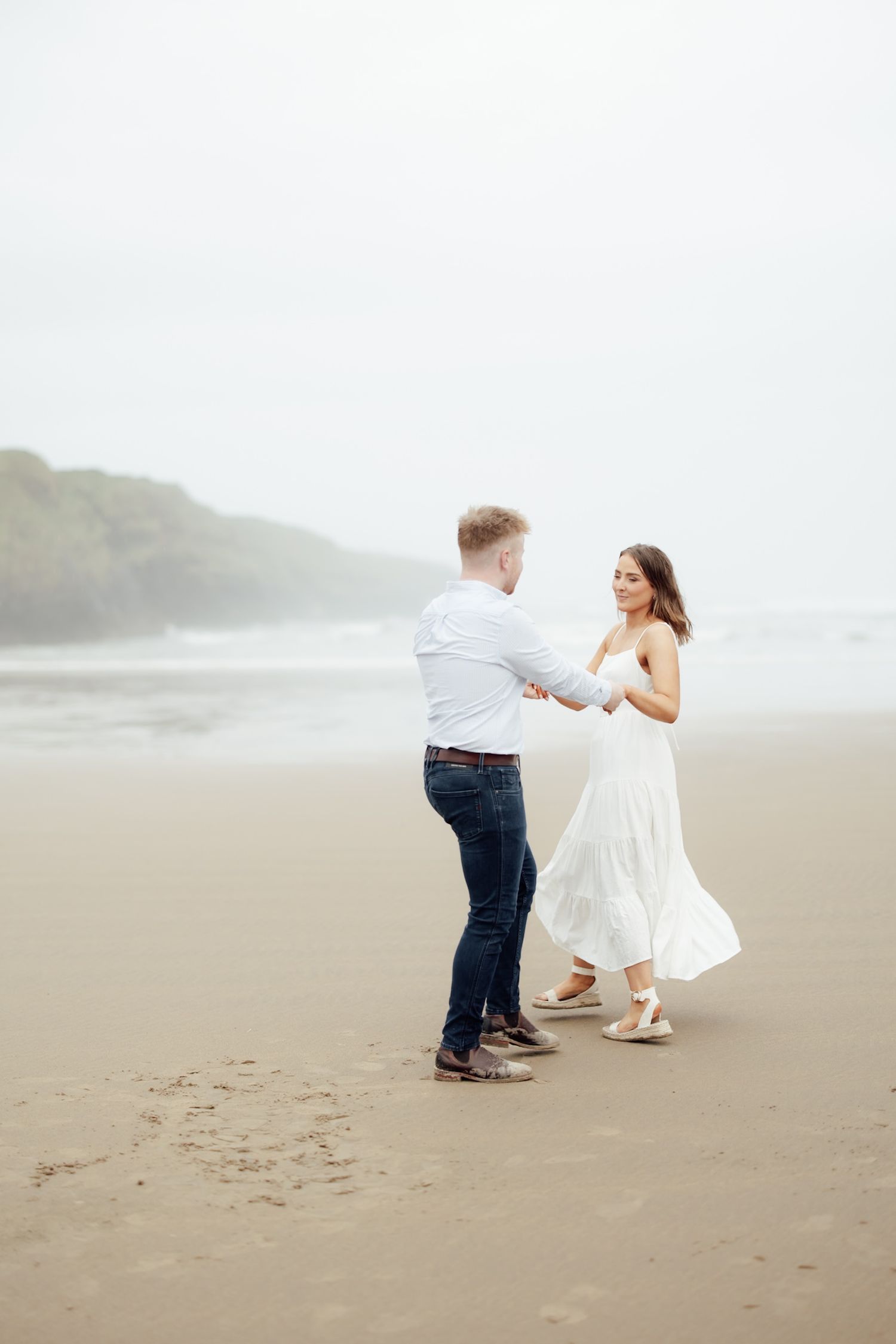 Couple dancing on a misty beach, woman in white dress, man in jeans and blue shirt, holding hands romantically.