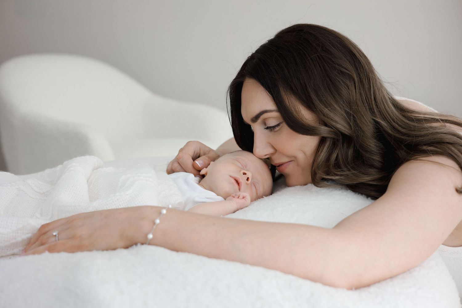 Mother lying on white bedding, gently nuzzling her sleeping newborn baby in a tender intimate portrait.