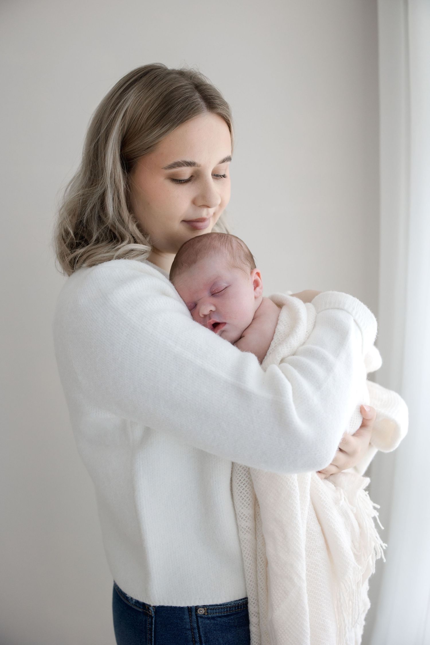 A young blonde mother in a white sweater cradles her sleeping newborn wrapped in a white blanket.