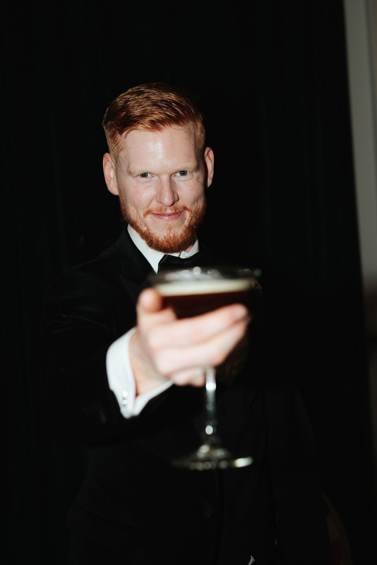 Red-bearded man in black tuxedo raising a dark cocktail glass toward camera against dark background.