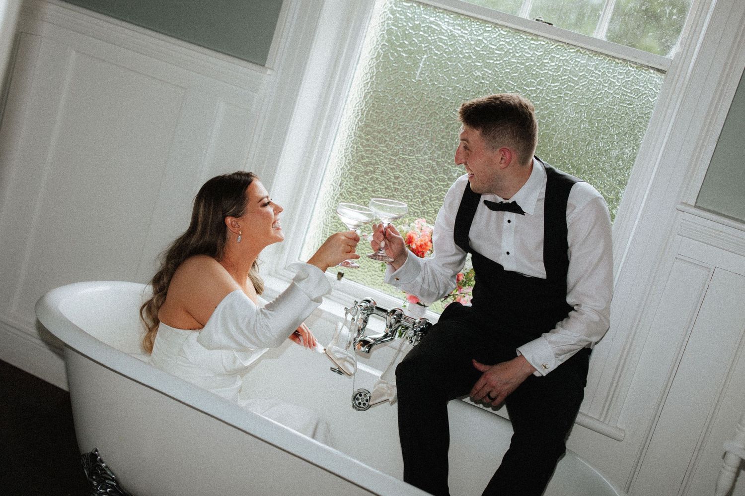 Bride and groom toasting champagne glasses in a clawfoot bathtub by a window in elegant wedding attire.