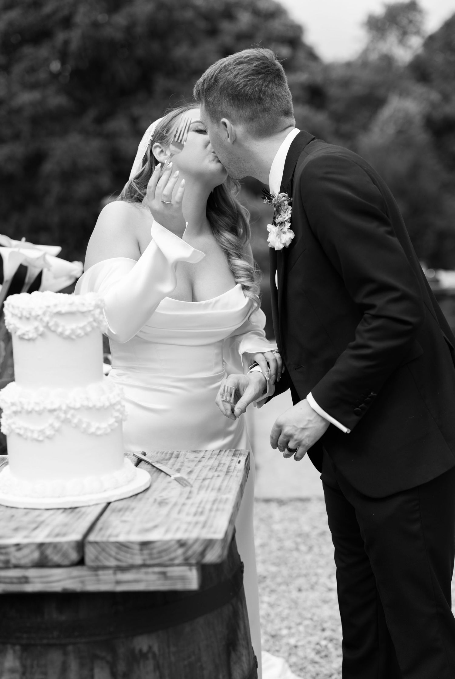 Black and white photo of bride and groom sharing a kiss while cutting their wedding cake outdoors.