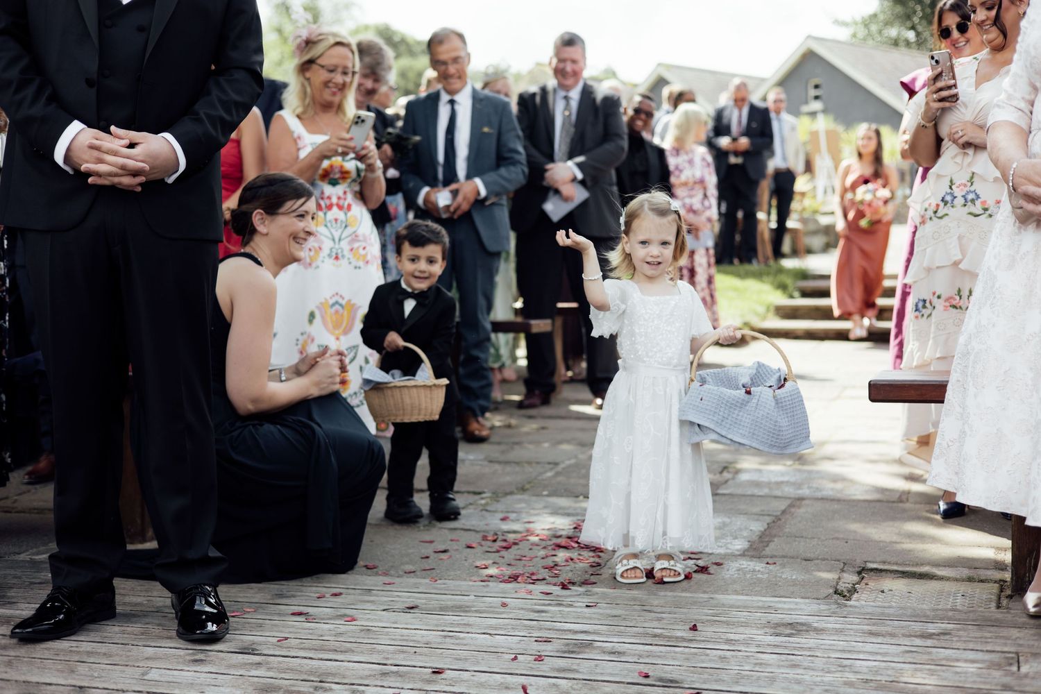 Adorable flower girl in white dress scattering petals on wooden deck during an outdoor wedding ceremony.