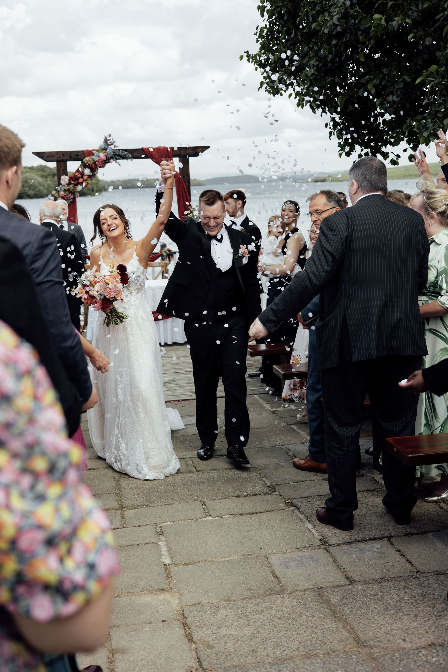 Bride and groom walk joyfully down the aisle as guests throw confetti at an outdoor waterside wedding ceremony.