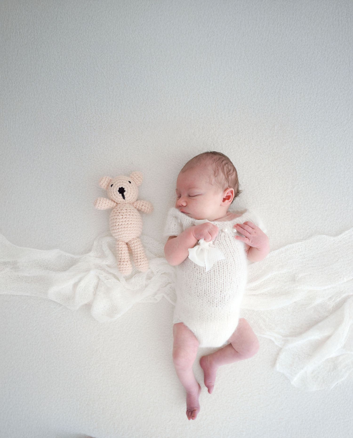 Sleeping newborn baby in white romper lying beside small crochet teddy bear on white fabric.