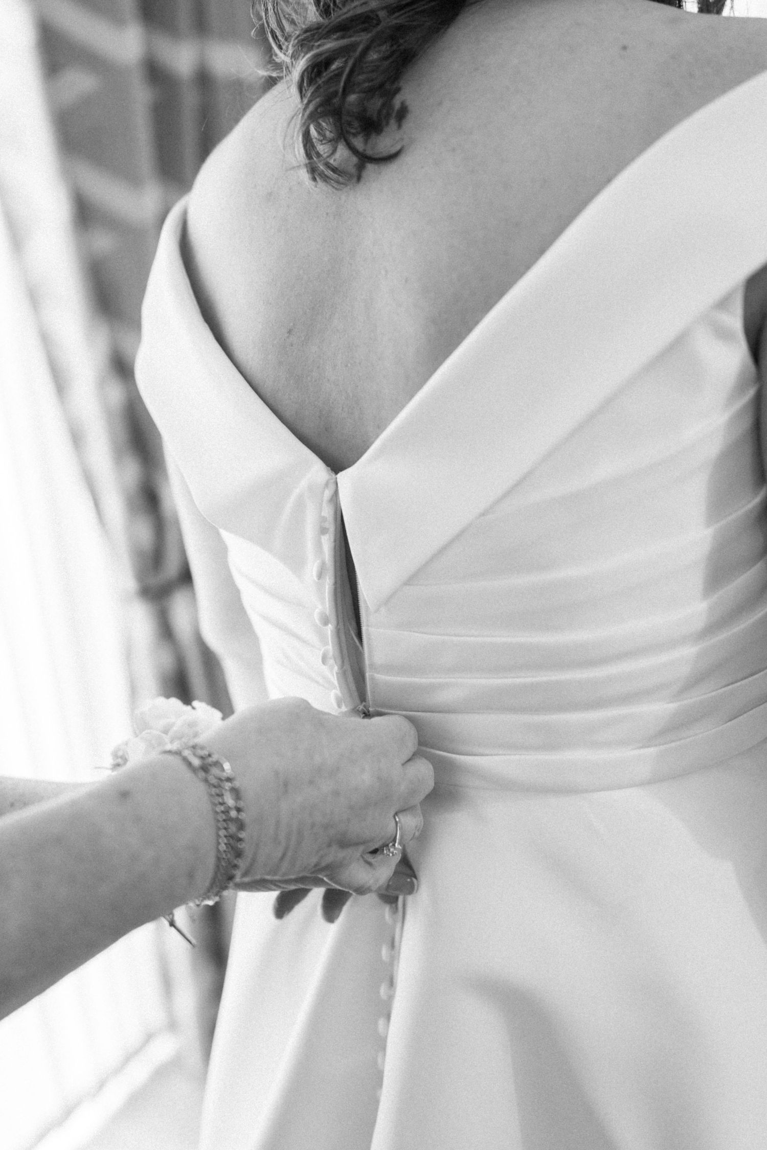 Black and white photo of a bride having her wedding dress buttoned up from behind by a helping hand.