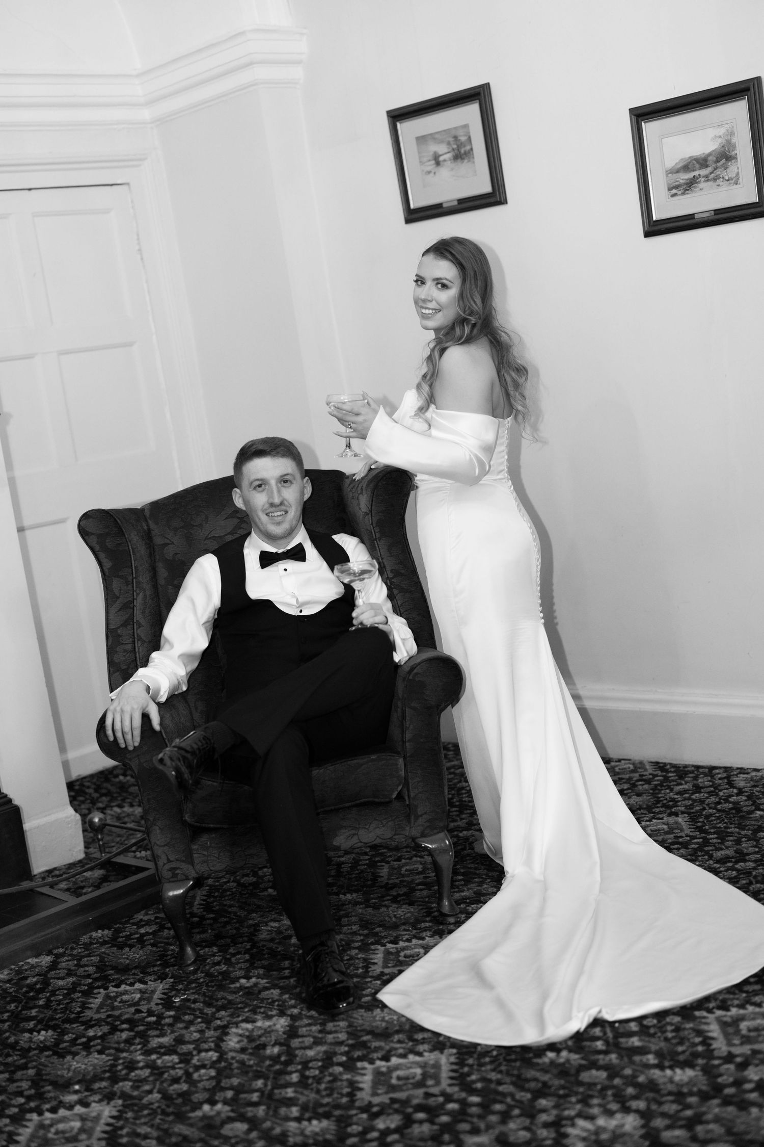 Black and white wedding photo of groom seated in armchair and bride standing beside him holding champagne glass.
