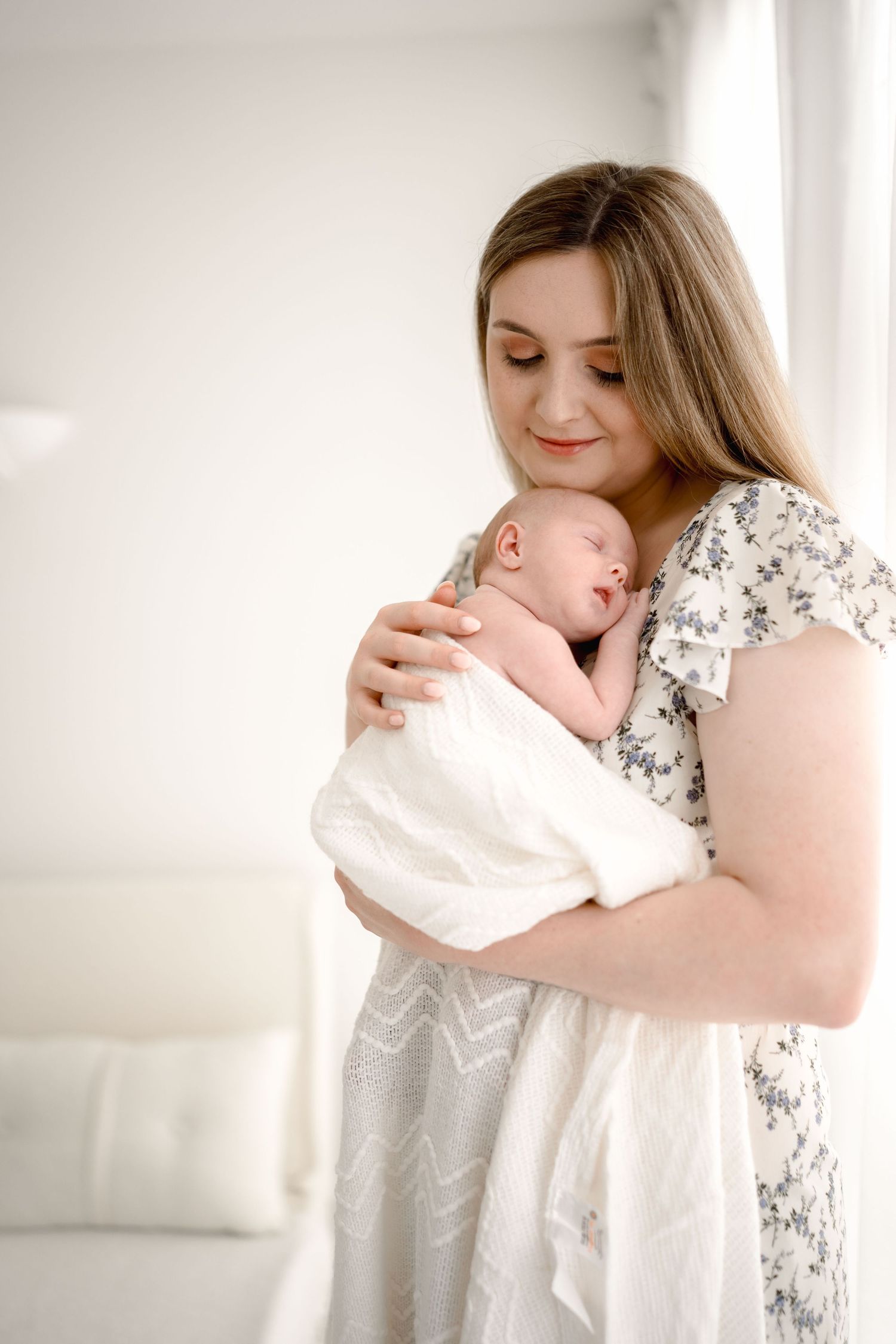 Young mother in a floral dress gently cradling her sleeping newborn baby in a bright white room.