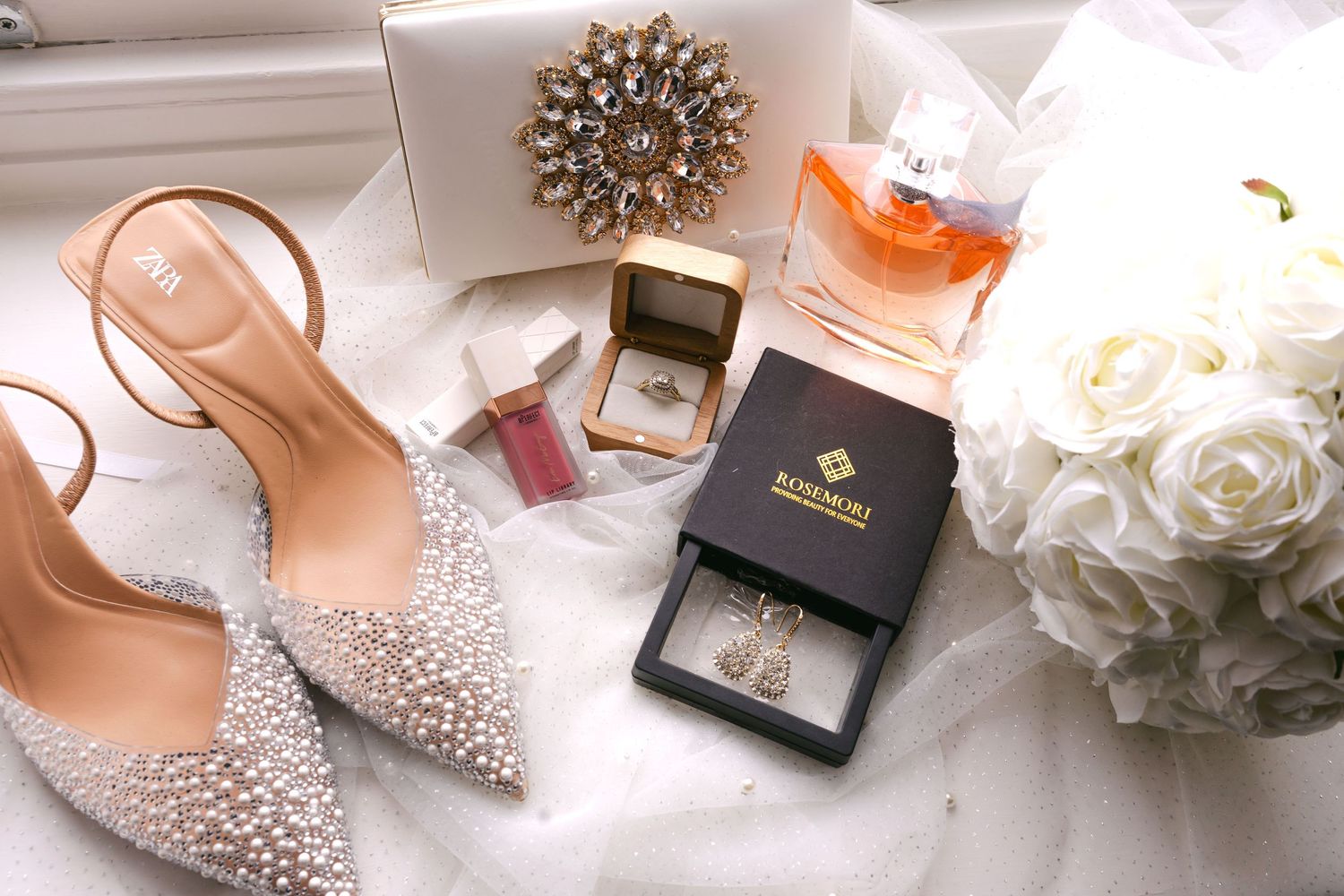 Wedding flat lay featuring pearl heels, diamond ring, gold brooch, perfume, earrings, and white roses bouquet.