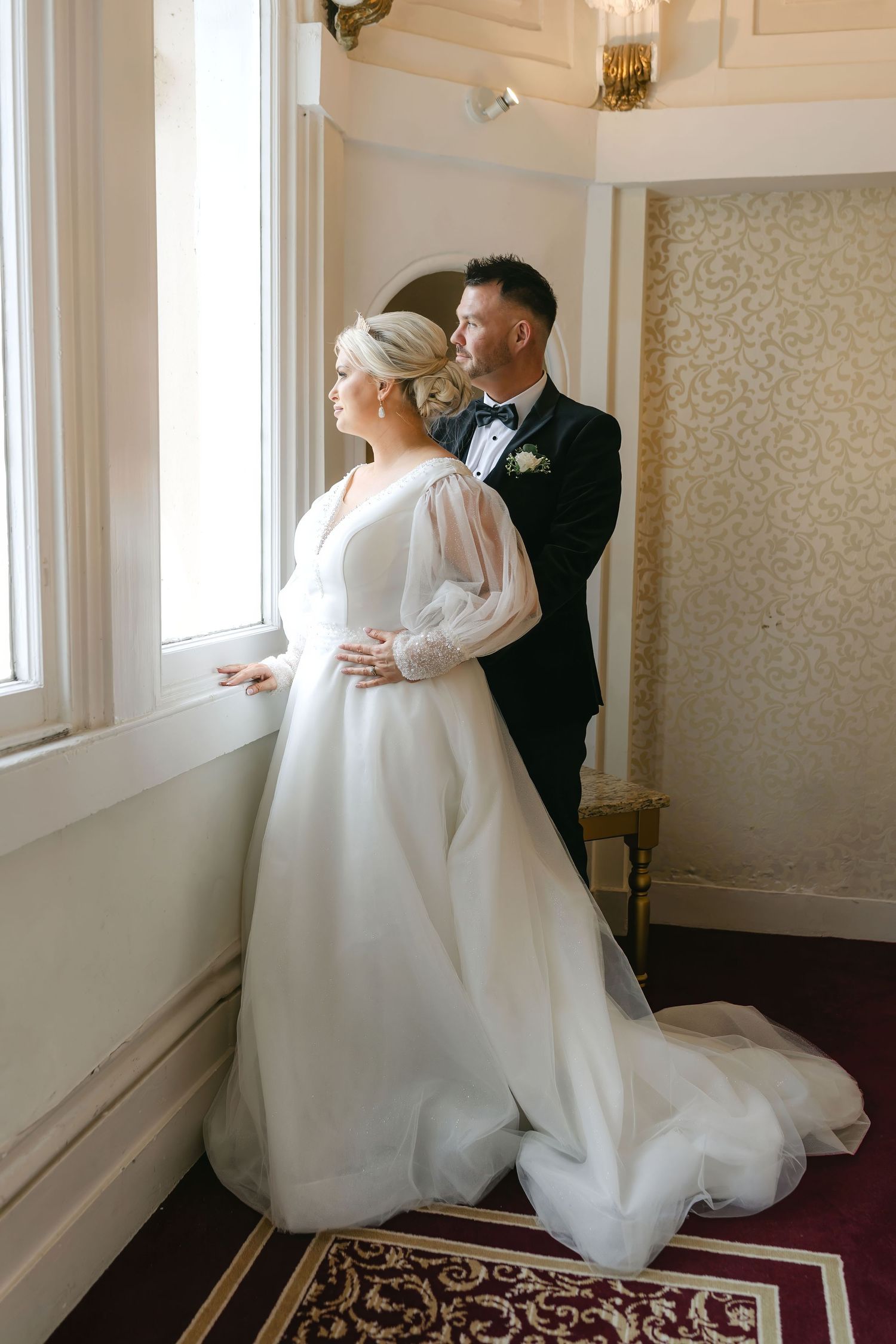 A bride in a white gown with sheer sleeves and groom in a tuxedo gaze out a window together on their wedding day.