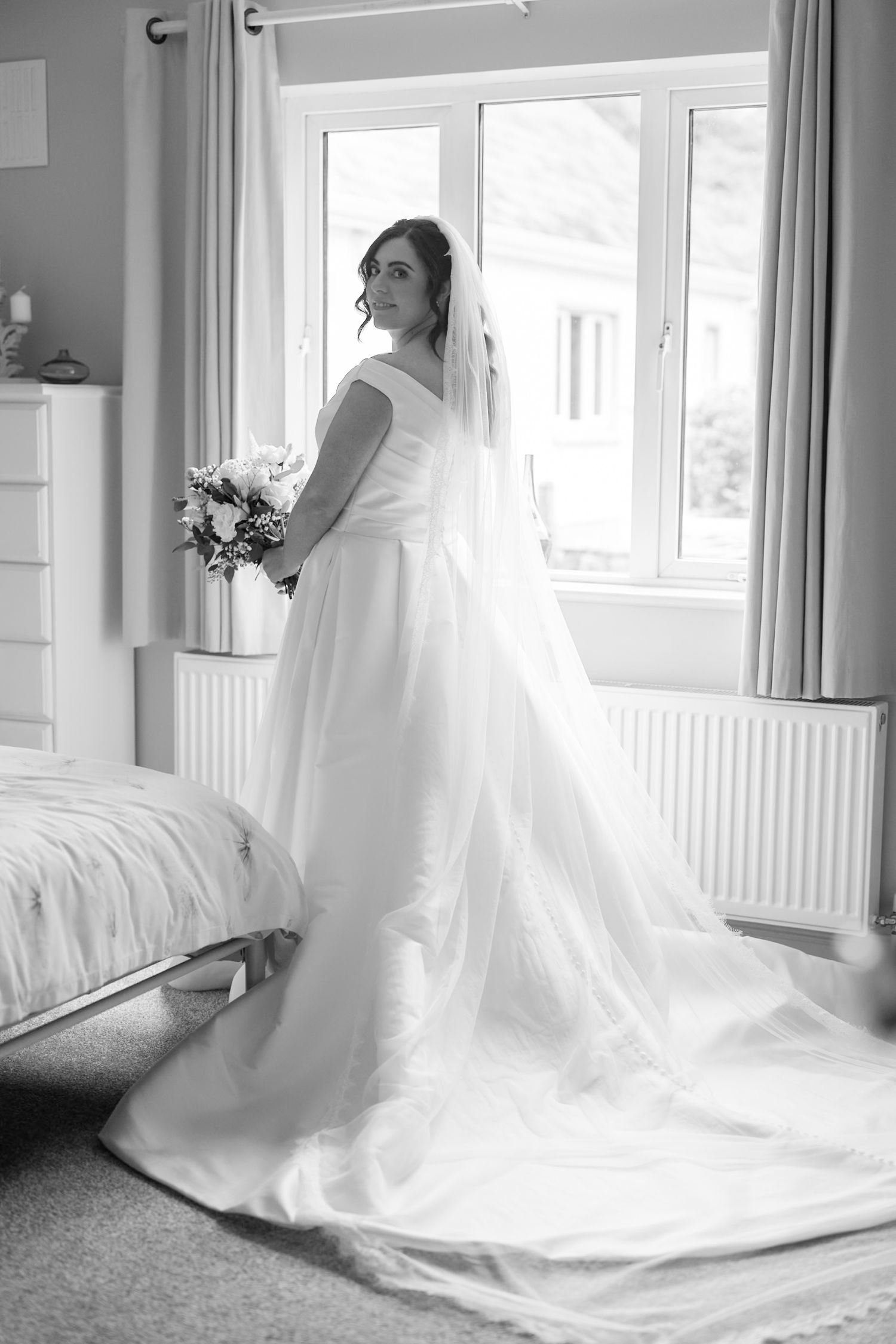 Black and white photo of a bride in a flowing wedding gown with veil, holding a bouquet by a sunlit bedroom window.