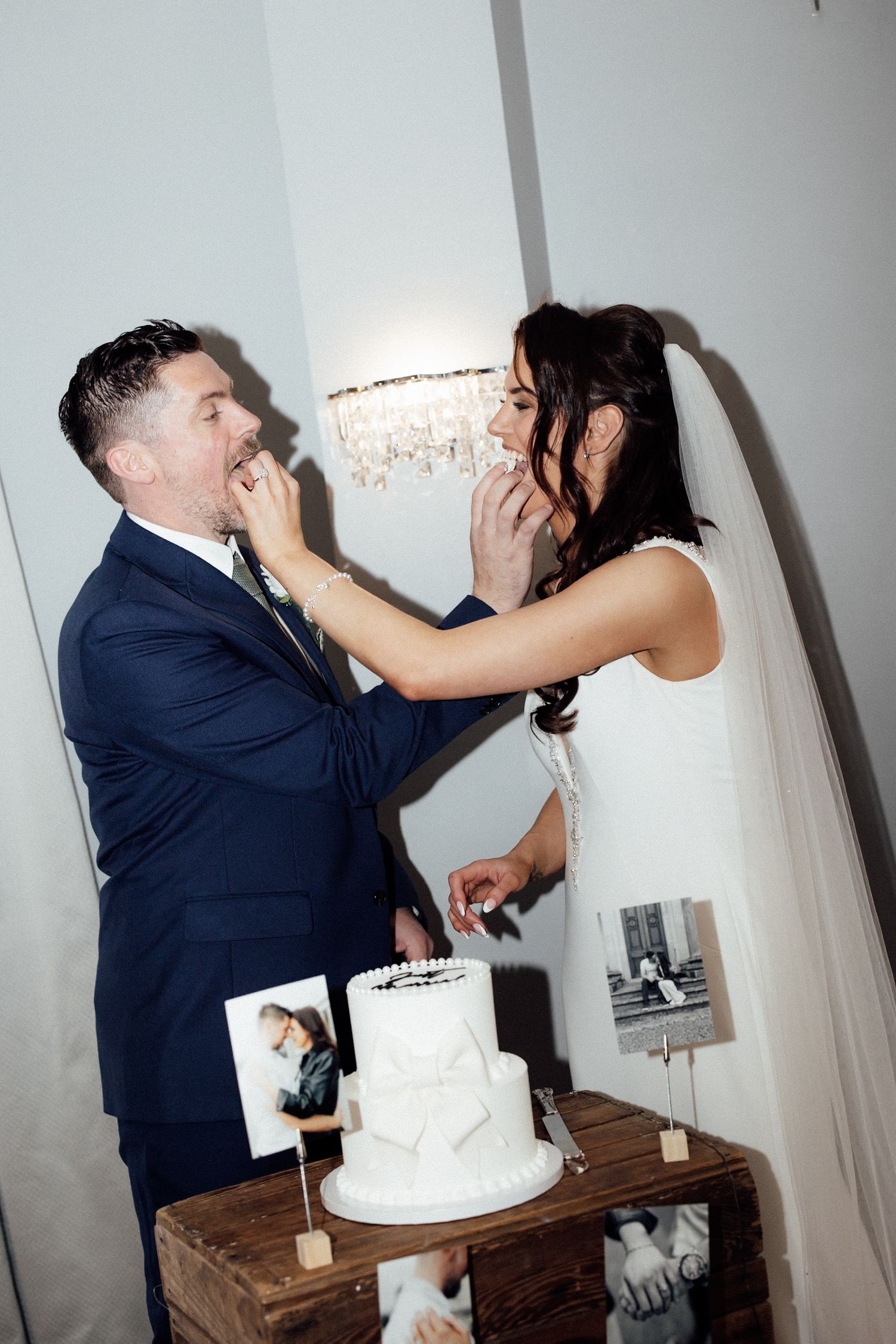 Bride and groom feeding each other wedding cake at elegant reception with white tiered cake and vintage photos on rustic wooden table.