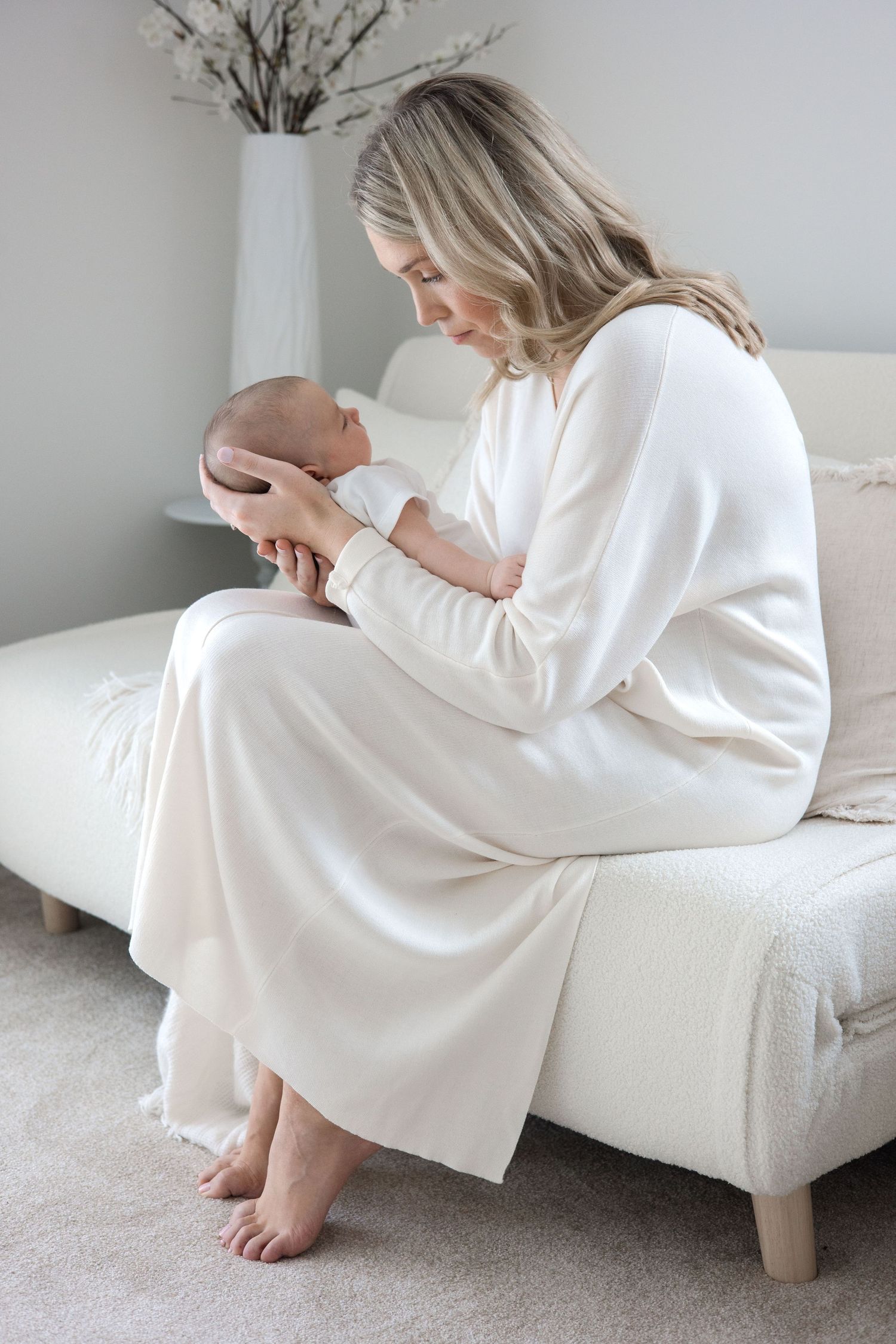 A blonde mother in a cream dress sits barefoot on a sofa, gazing at her newborn baby.