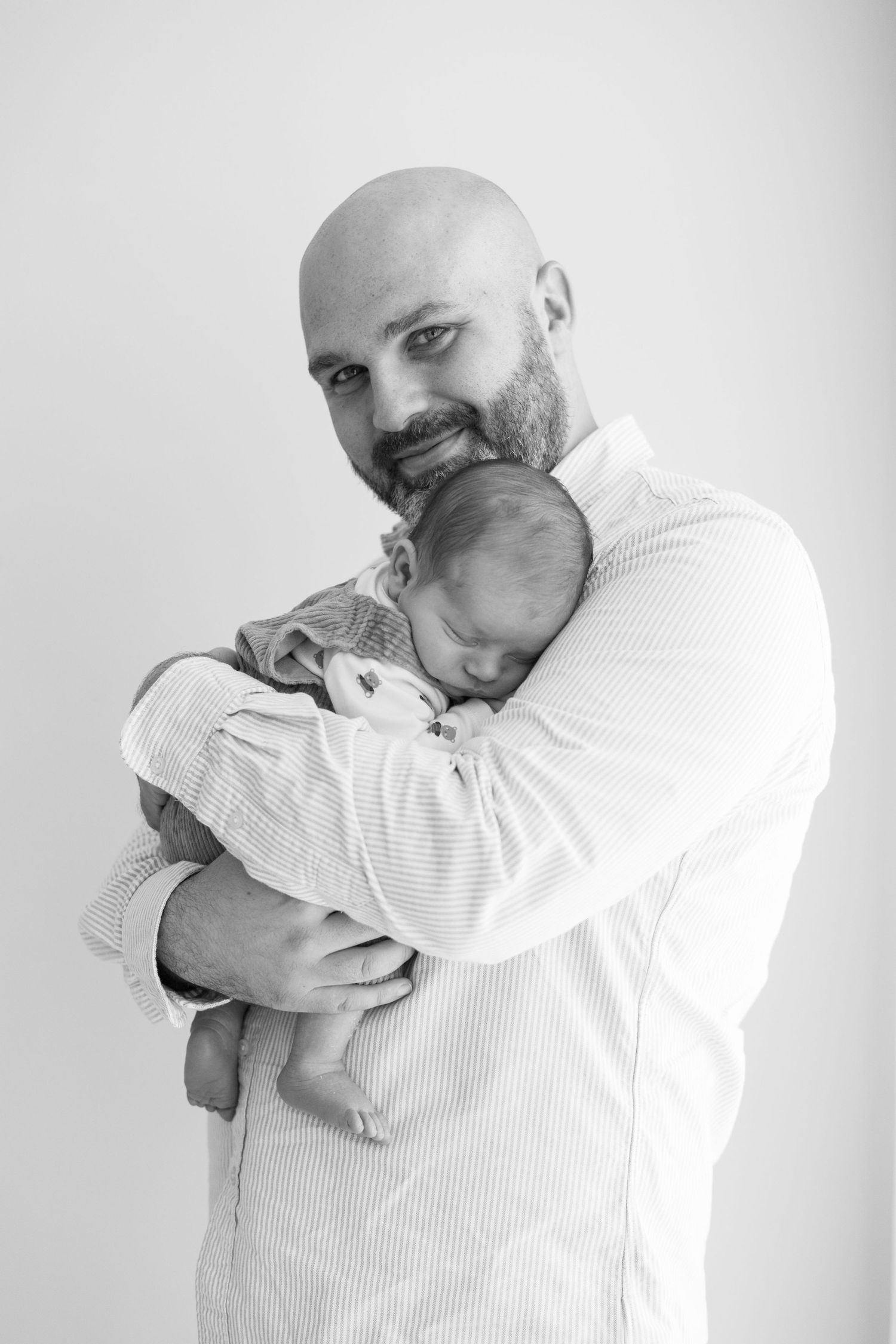 Black and white portrait of bald bearded father smiling while holding sleeping newborn.