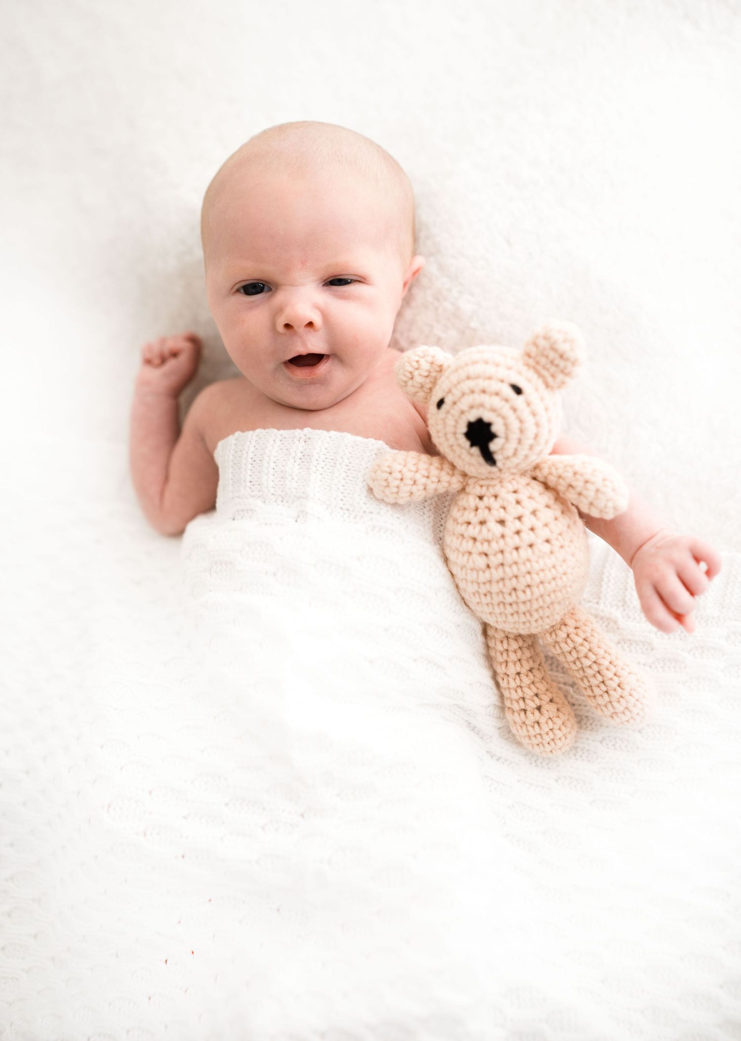Awake newborn baby lying on white blanket beside a small crocheted teddy bear, looking at camera.