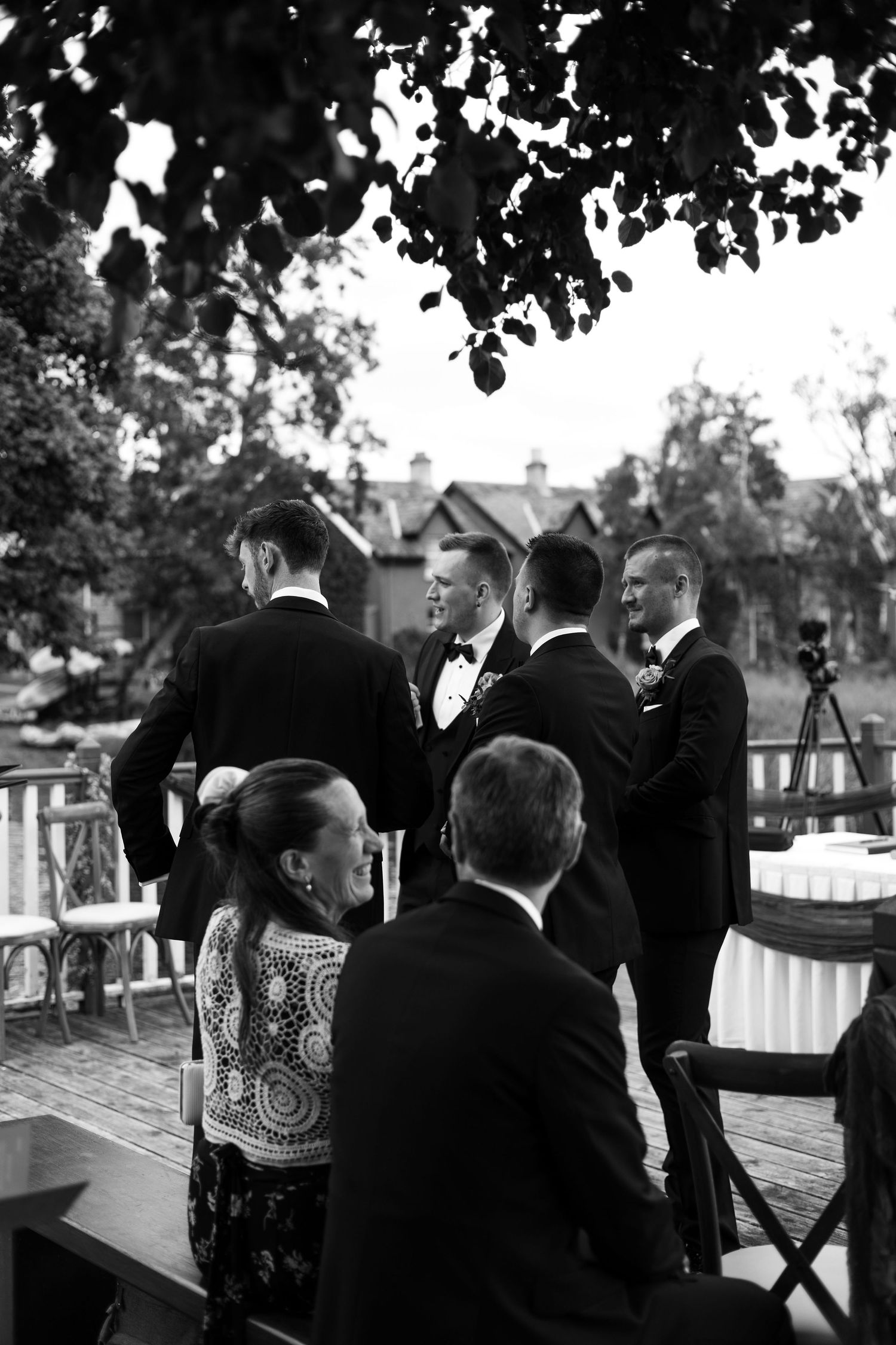 Black and white photo of groomsmen in tuxedos gathered at an outdoor wedding ceremony on a wooden deck.
