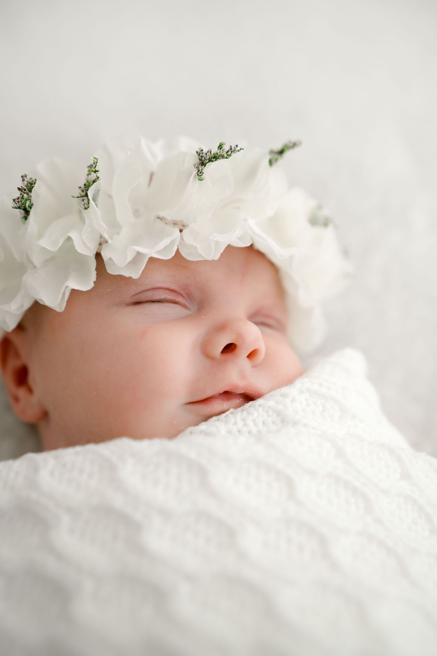 Sleeping newborn baby wearing a white floral crown, swaddled in a white knit blanket.