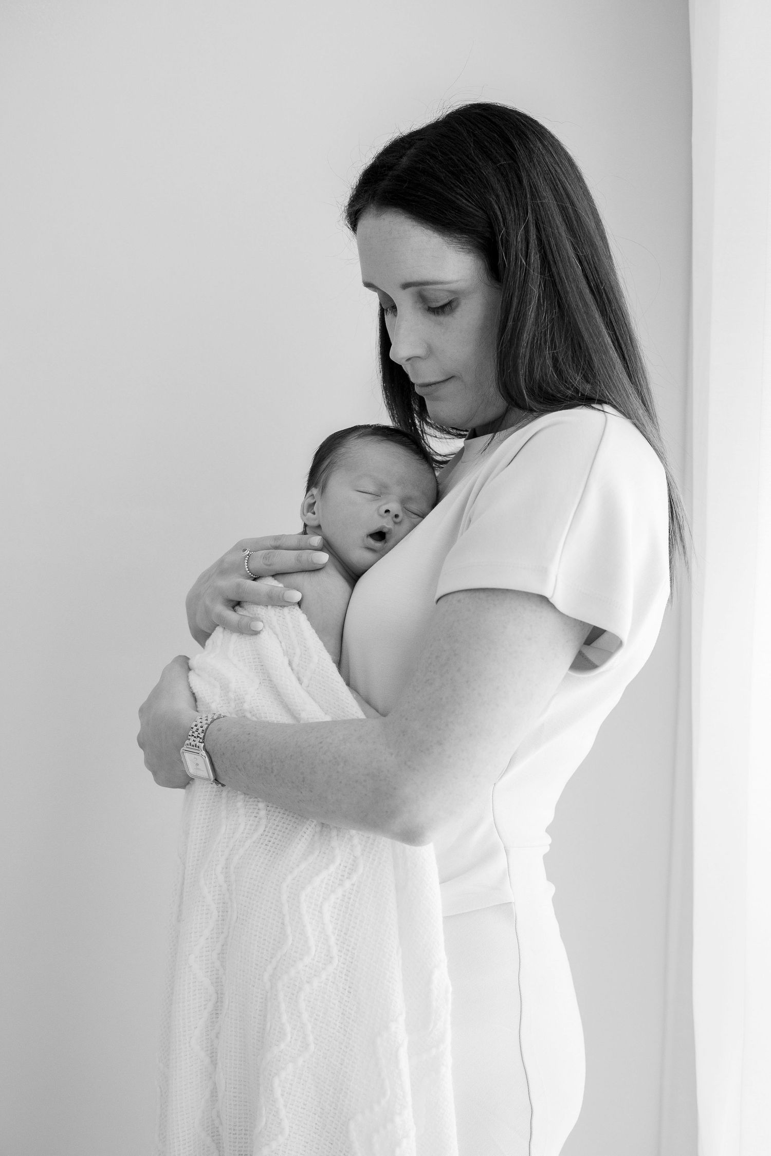 A dark-haired mother in a white dress holds her sleeping swaddled newborn in black and white.