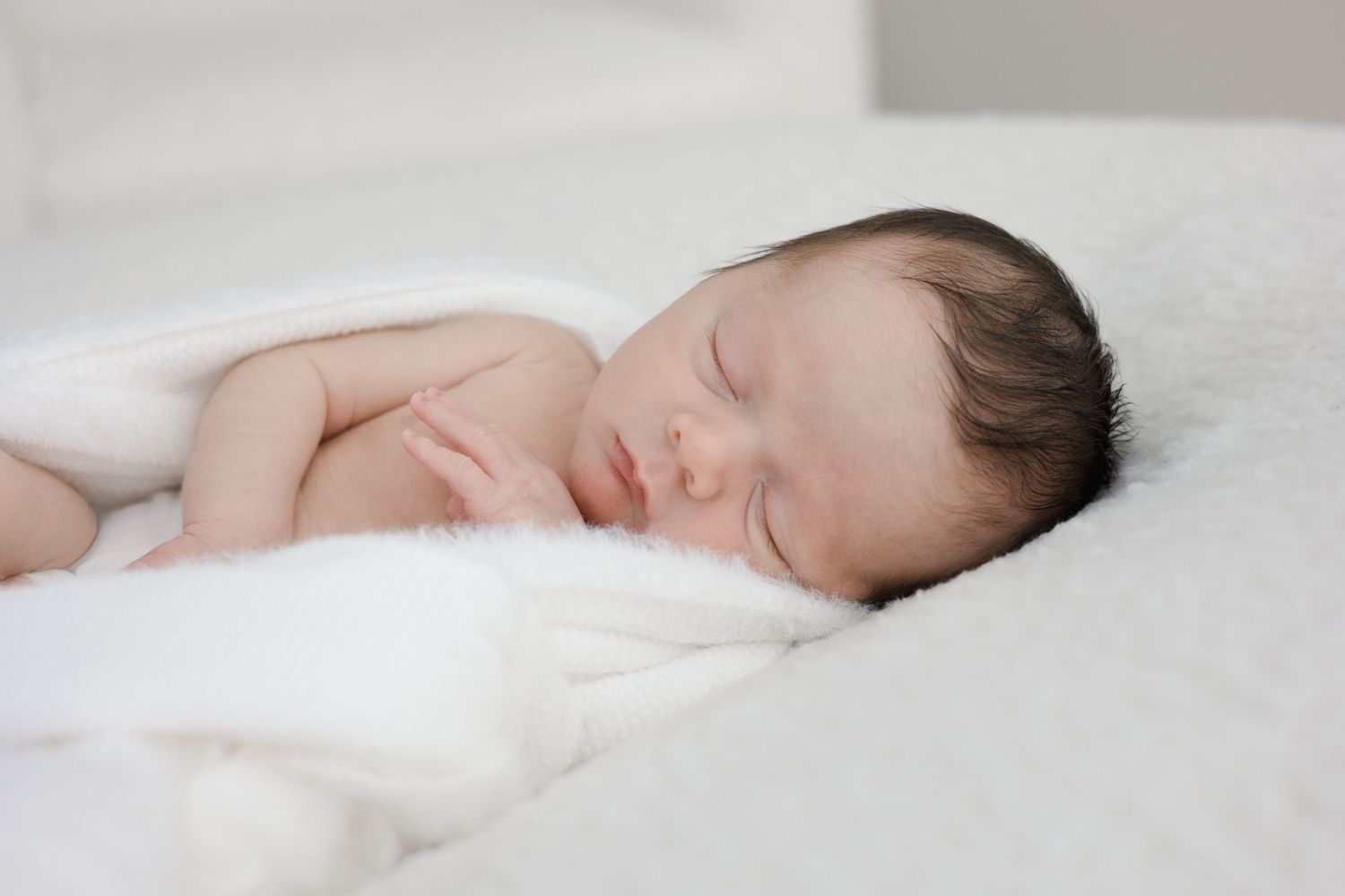 Peaceful newborn baby with dark hair sleeping on a white blanket.