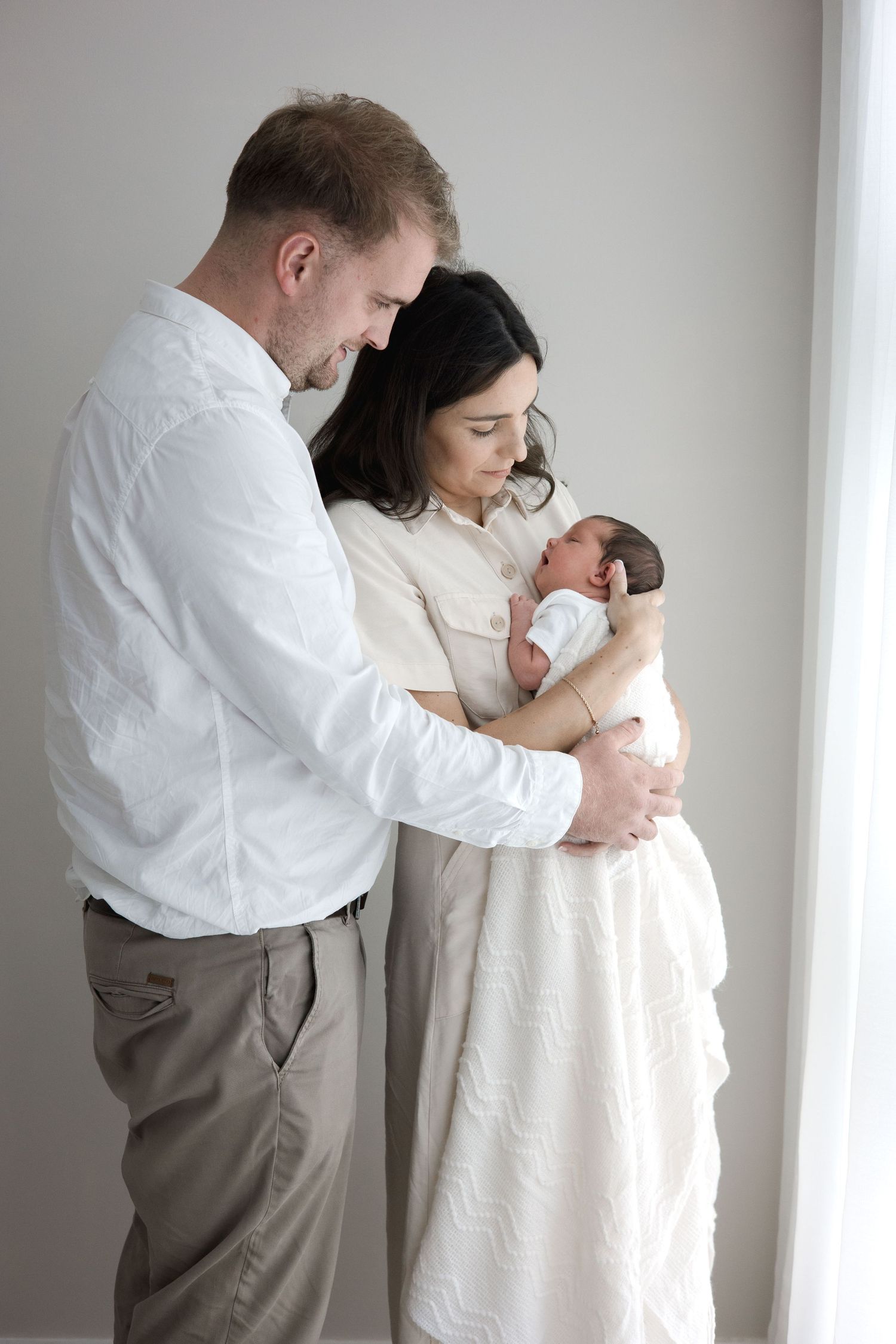 New parents in white outfits standing by window together cradling their newborn baby.