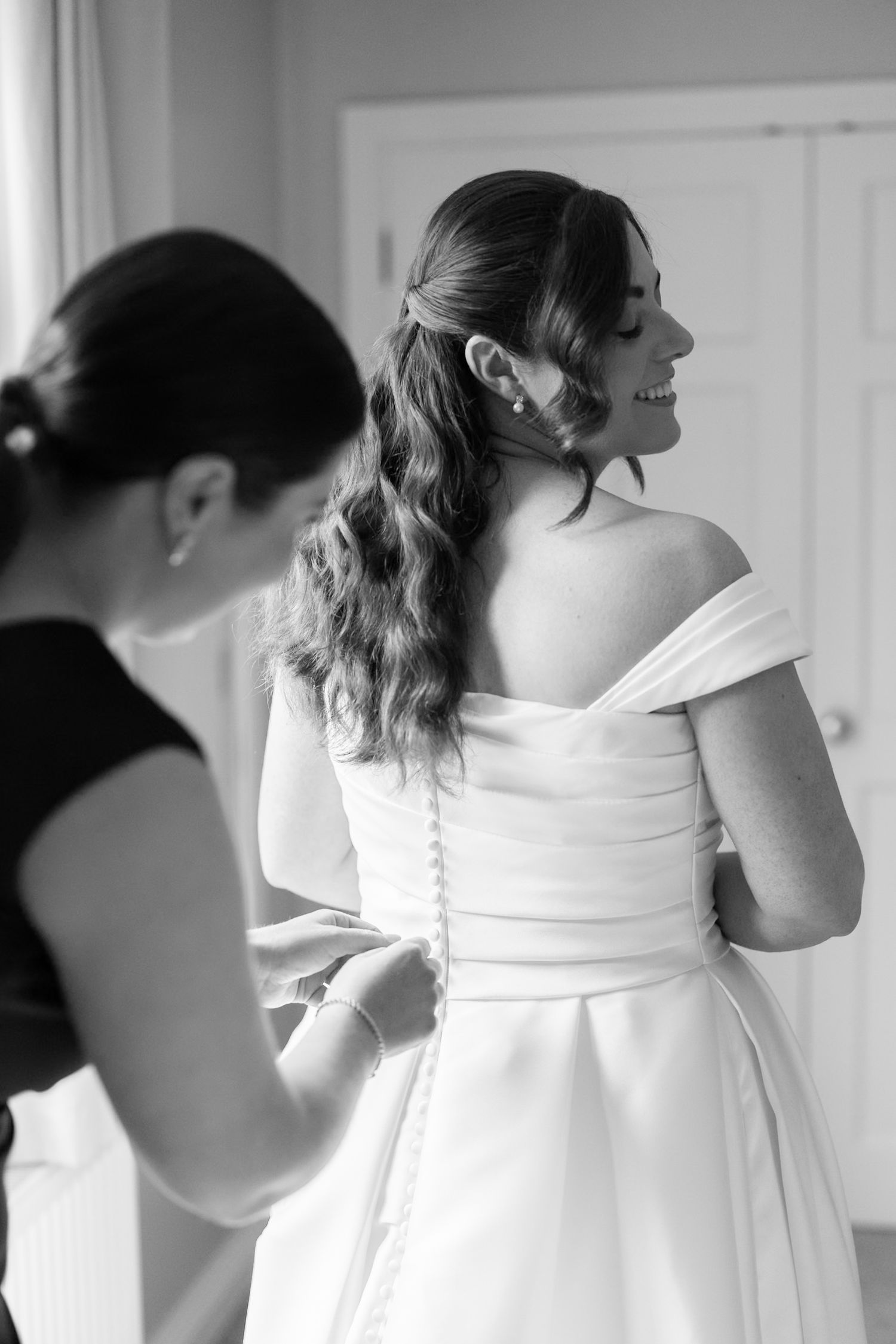 Black and white photo of a bride smiling as her bridesmaid buttons up her elegant off-shoulder wedding gown.