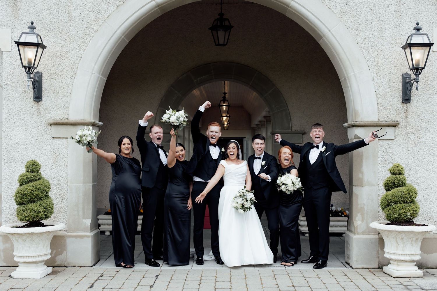 Wedding party celebrates under stone arch, bride in white gown with bouquet, groom and groomsmen in tuxedos, bridesmaids in black.