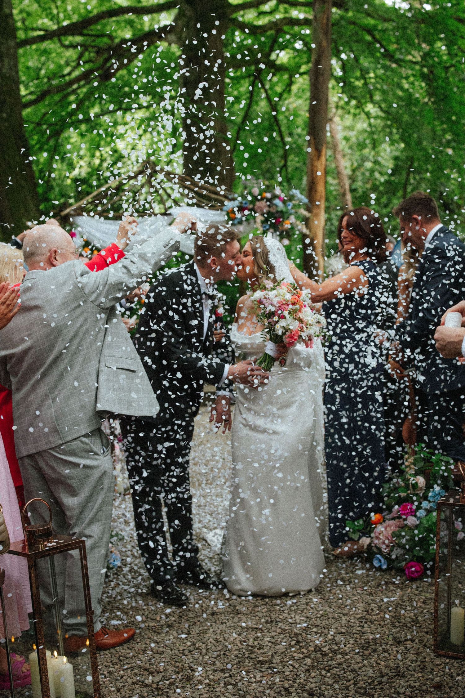 Bride and groom kissing while guests throw white confetti outdoors in a lush green woodland wedding setting.