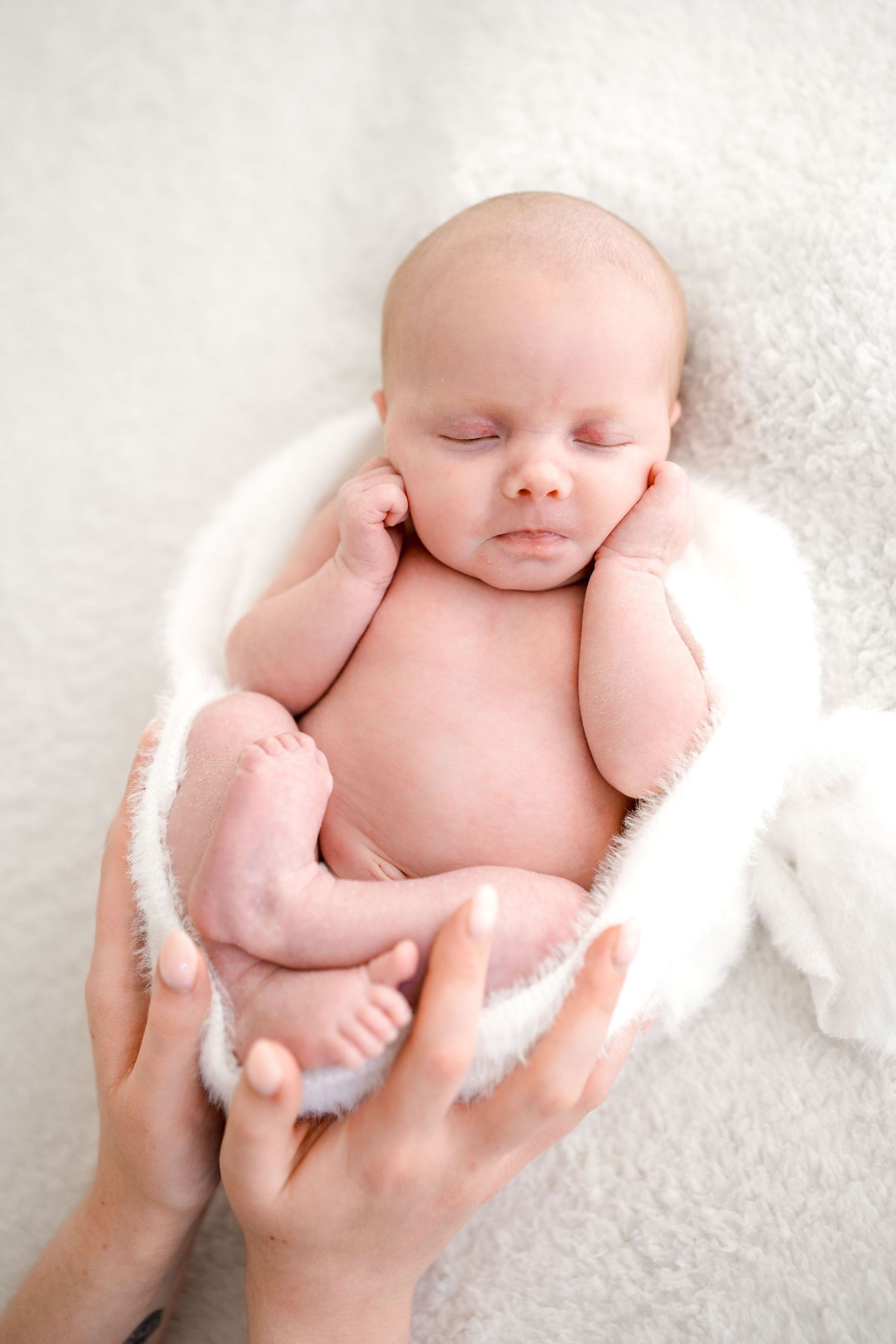 Mother's hands gently cradling a sleeping newborn baby wrapped in a soft white fluffy blanket.