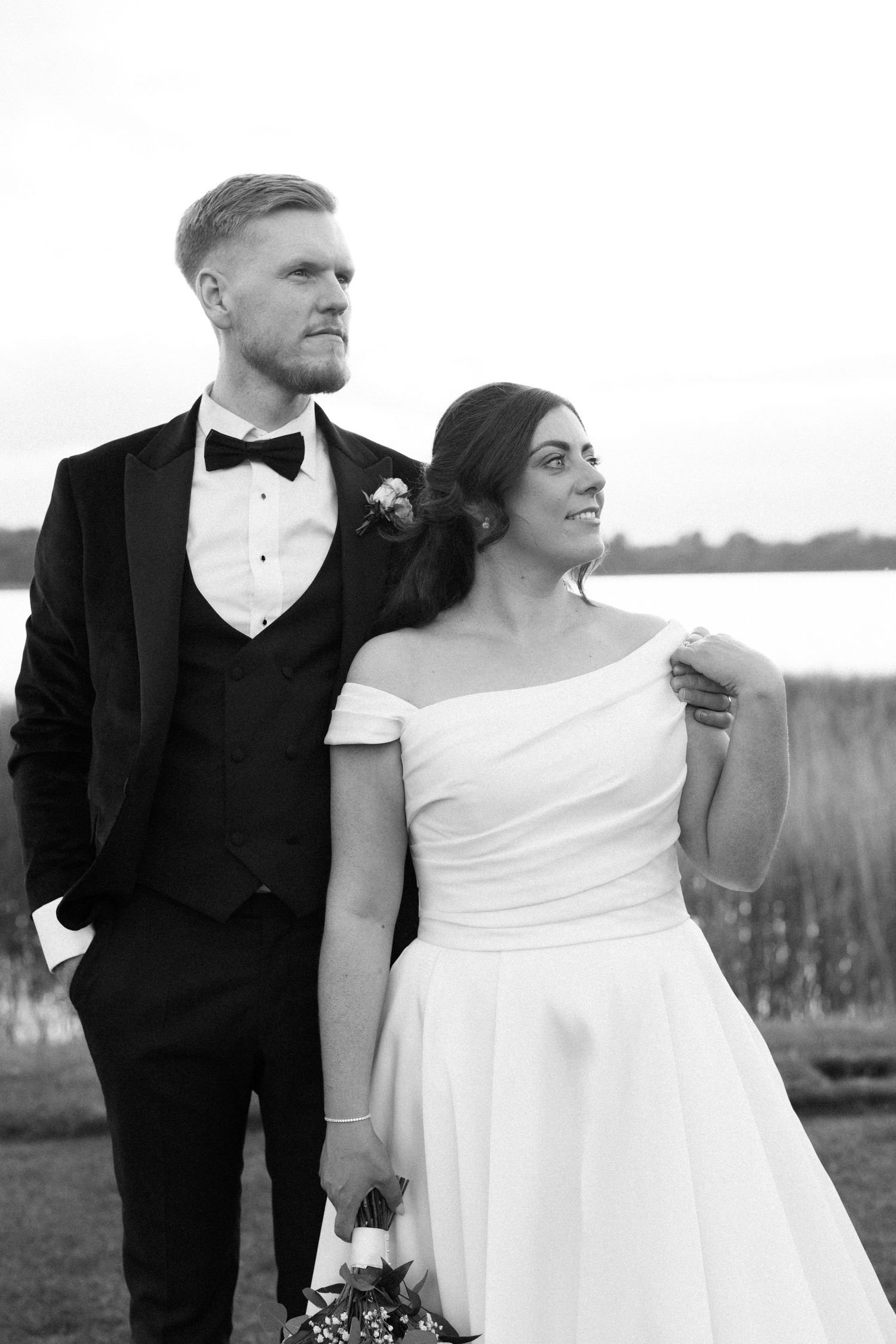 Black and white photo of a bride and groom posing outdoors near a lake, bride in off-shoulder gown holding bouquet.