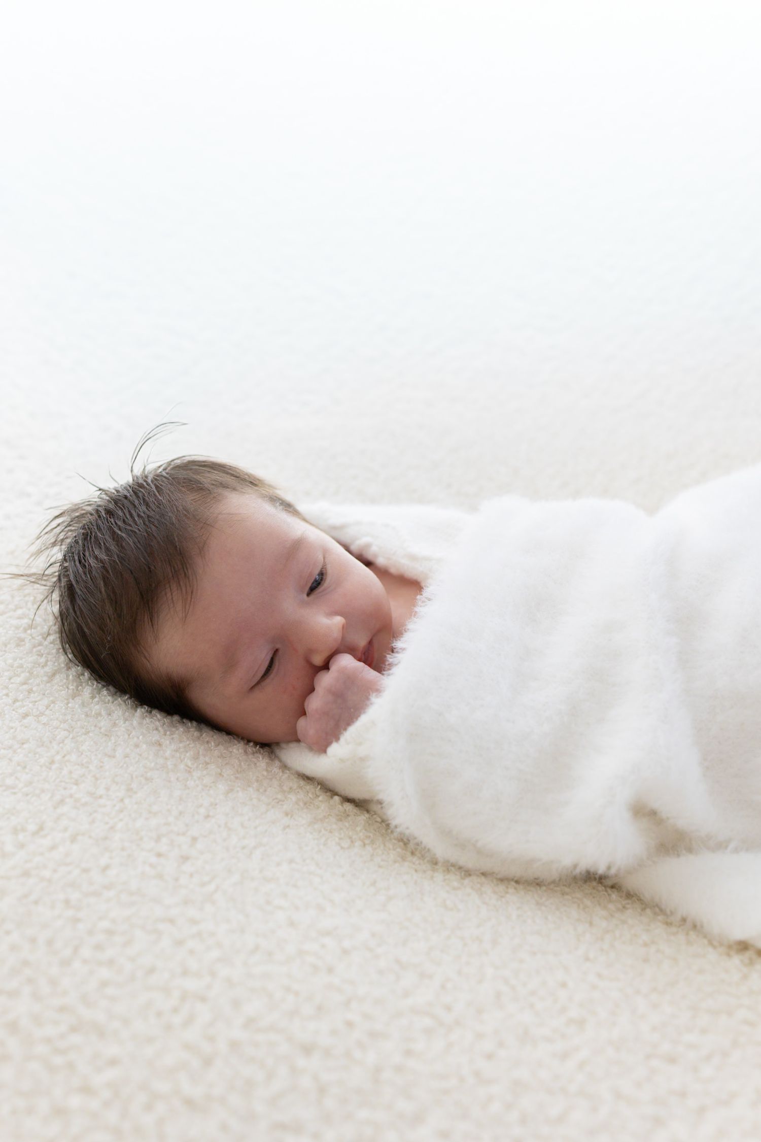 Peaceful newborn baby swaddled in white blanket lying on soft carpet.
