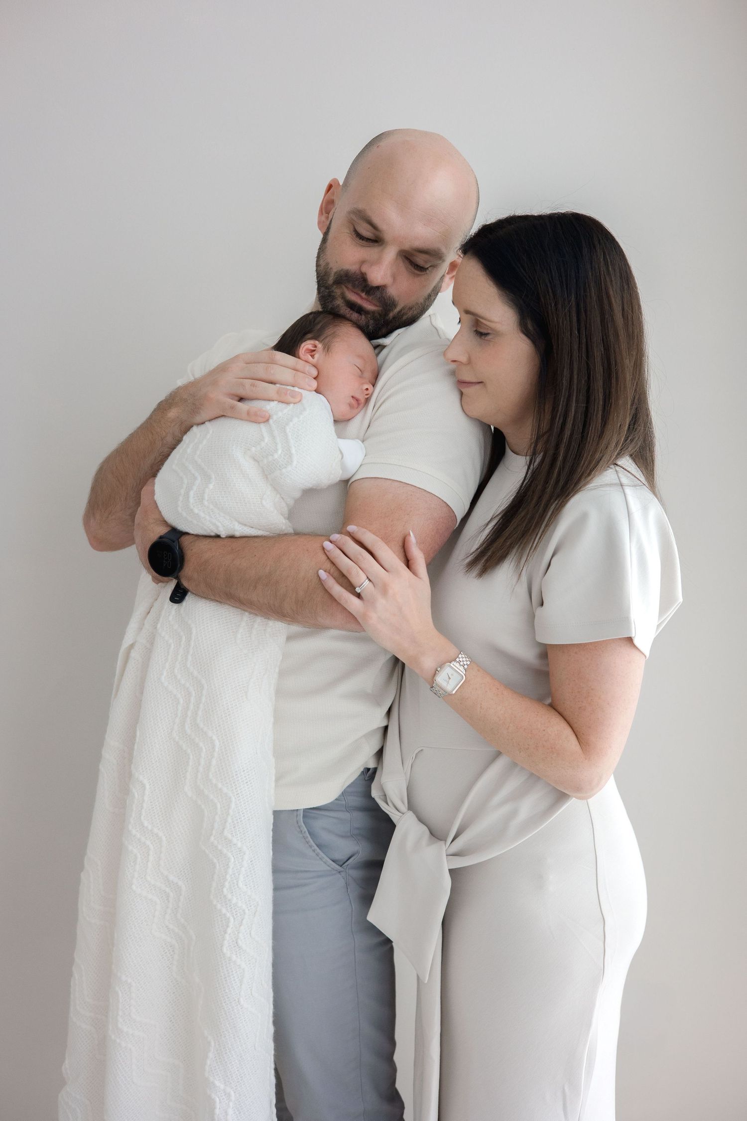 New parents in white outfits tenderly hold their swaddled newborn baby together against a light background.