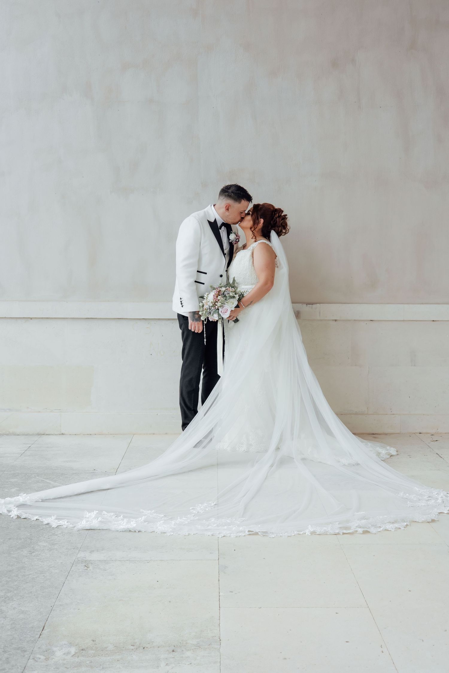 Bride and groom share a kiss in elegant wedding attire, with a flowing white cathedral veil trailing on marble floor.