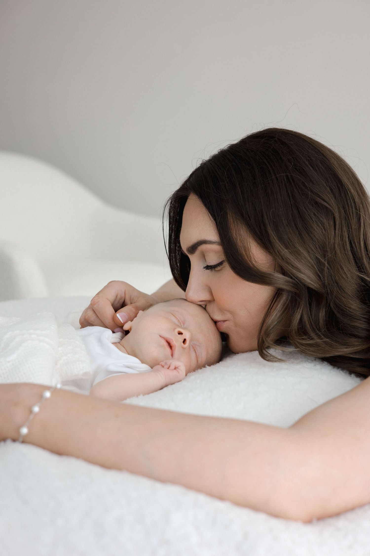 A dark-haired mother gently kisses her sleeping newborn baby lying on a soft white surface.